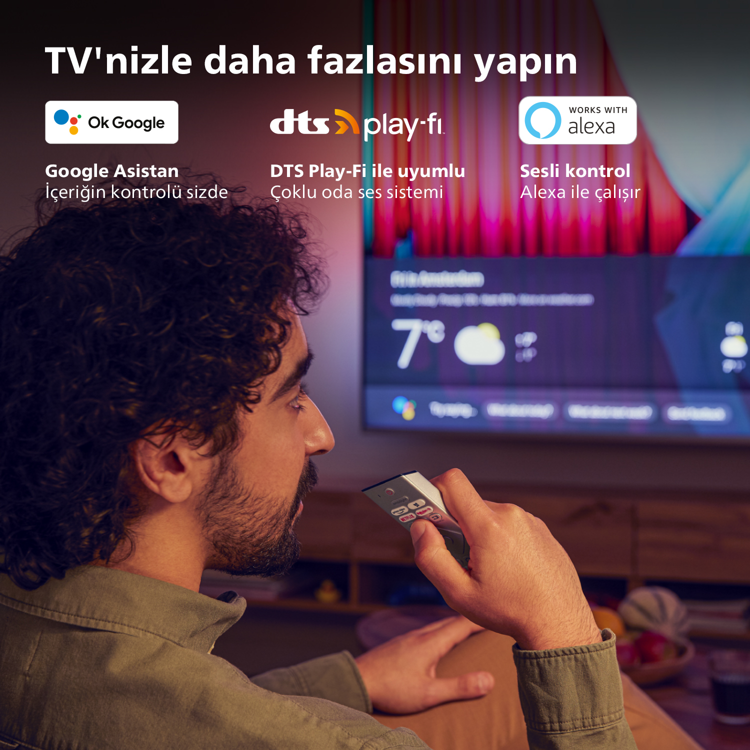 Elinde kumanda olan adam, ekranda Google Asistan, DTS Play-Fi ve Alexa logolarıyla TV izliyor.
