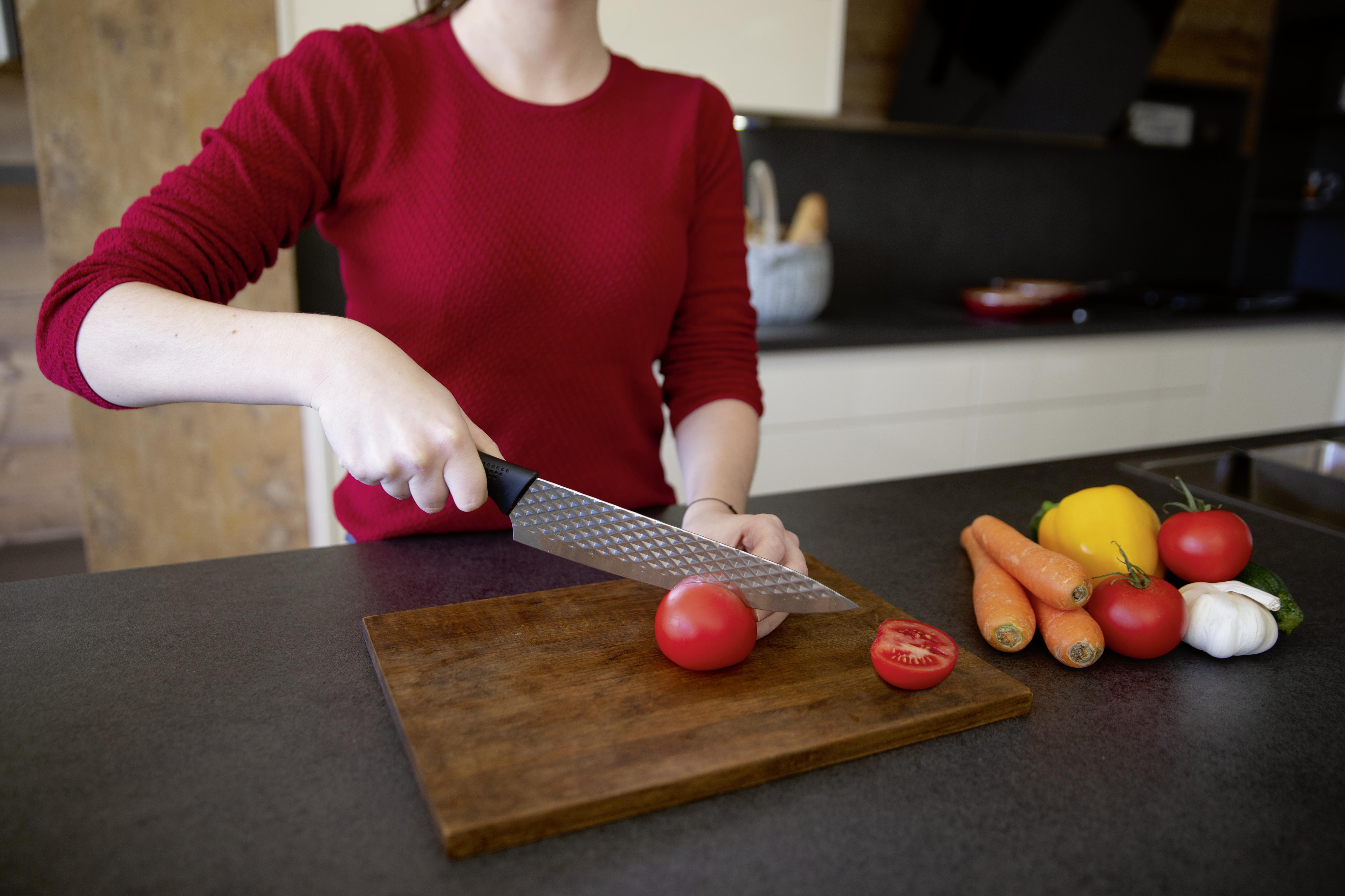 Eine Person schneidet eine Tomate auf einem Holzbrett mit einem Messer. Gemüse wie Karotten und Paprika sind daneben.