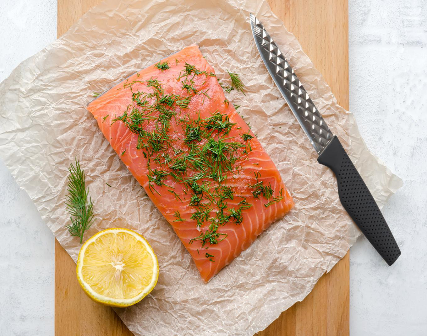 Ein Lachfilet mit Dill auf Pergamentpapier, eine Zitronenhälfte und ein Messer liegen auf einem Holzbrett auf weißem Untergrund.
