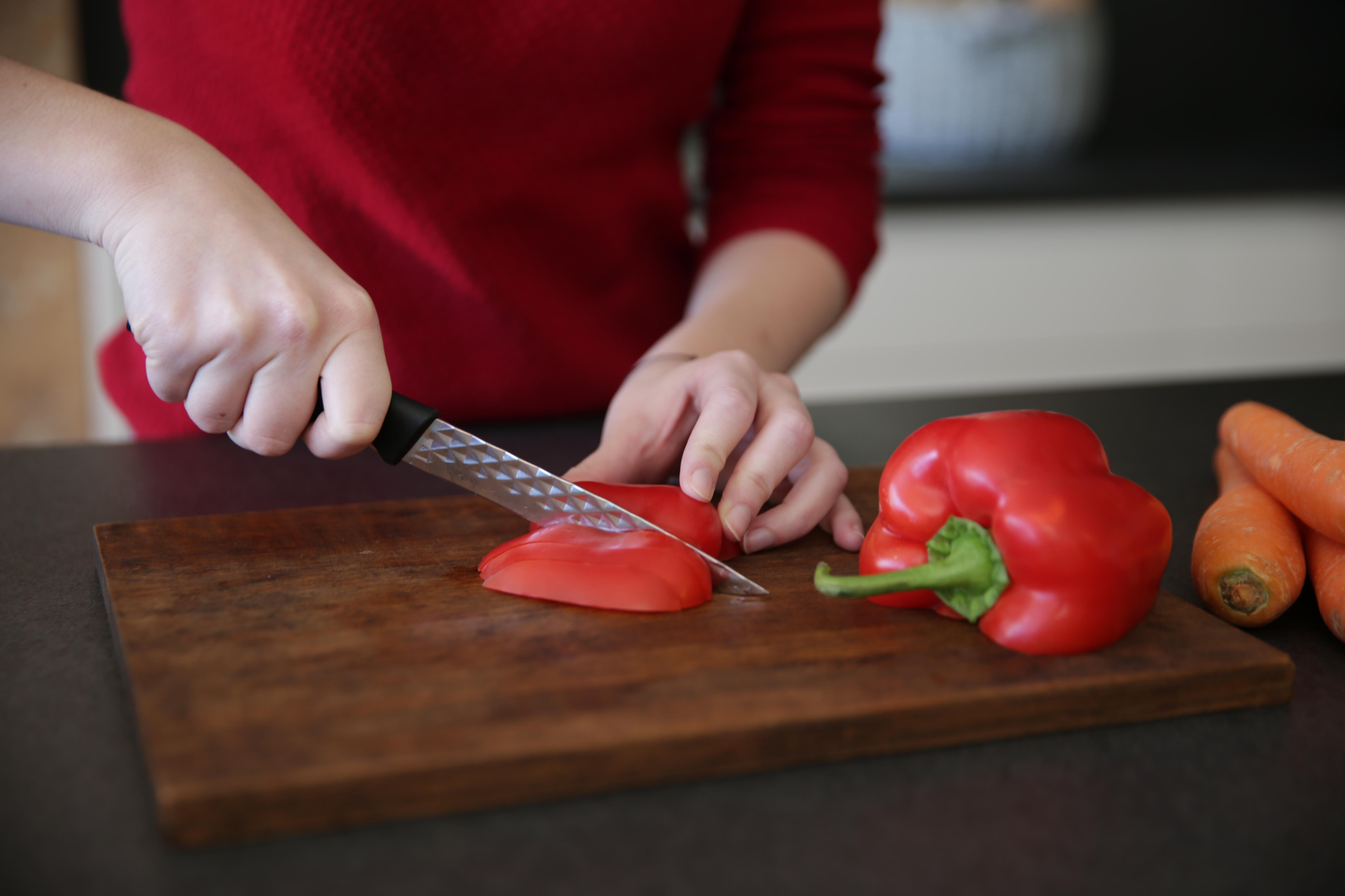 Eine Person schneidet eine rote Paprika auf einem Holzbrett mit einem Messer. Karotten und weitere Paprika liegen daneben.
