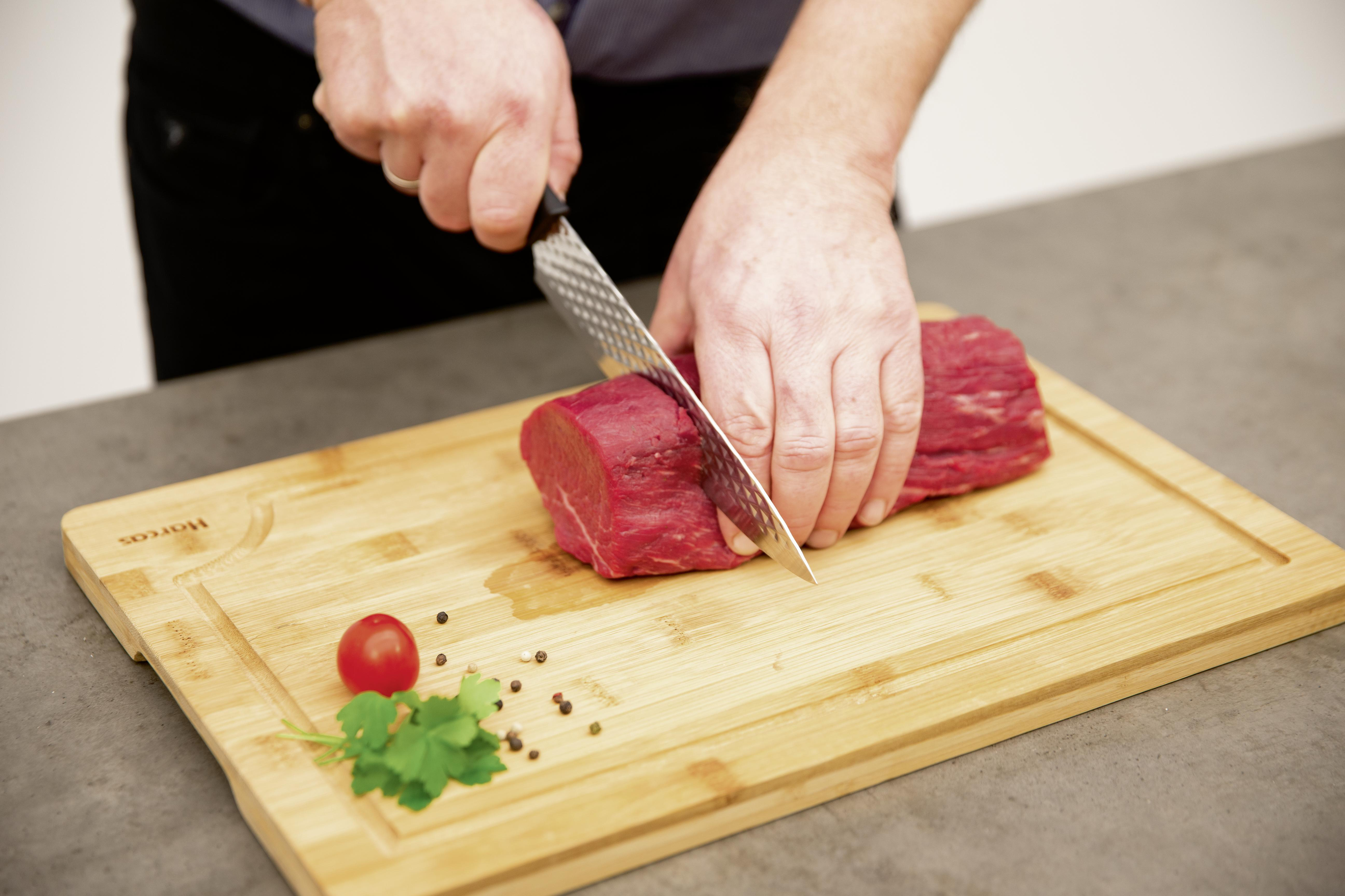Eine Person schneidet rohes rotes Fleisch auf einem Holzbrett. Daneben liegen Gewürze und eine Tomate.