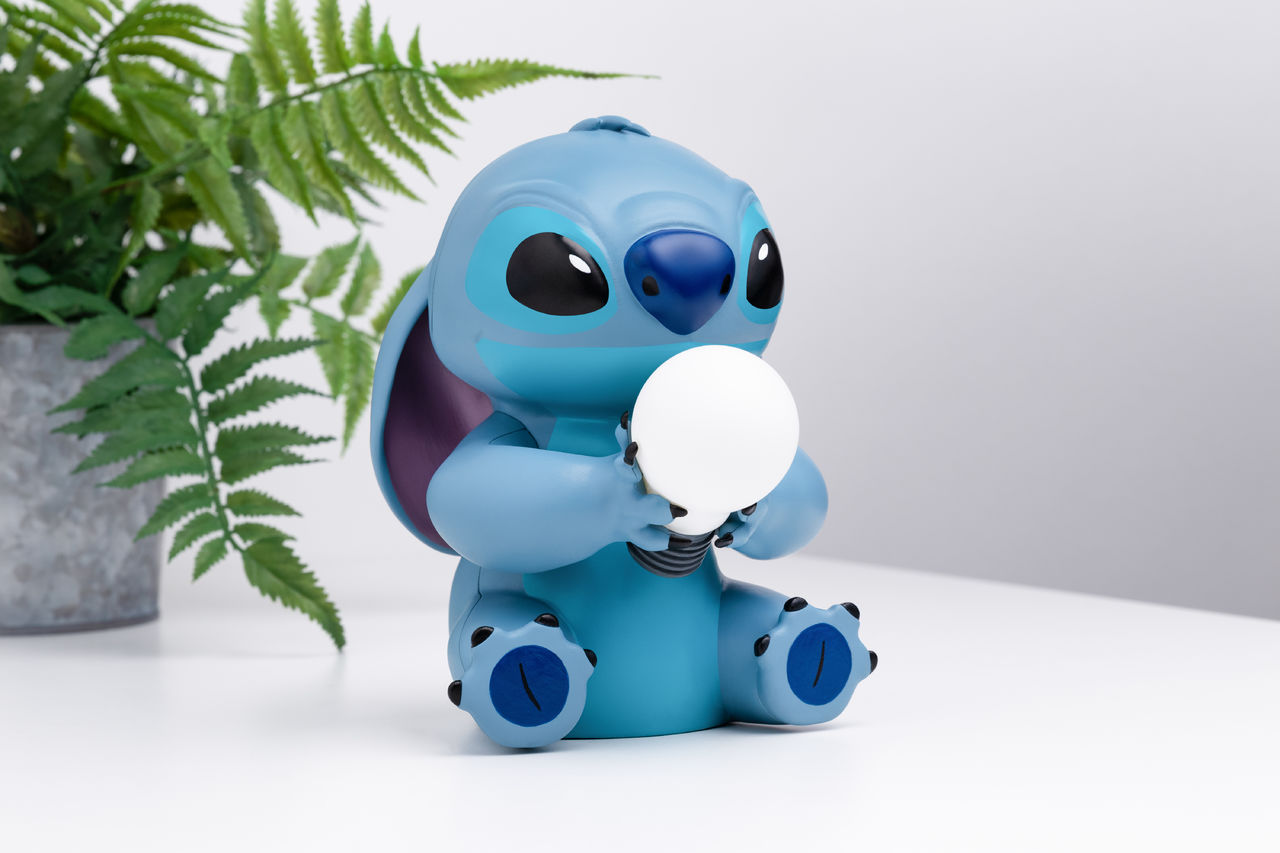 Stitch-Lampe, die eine Glühbirne hält. Er ist blau, sitzt auf weißer Oberfläche, Pflanze hinten.