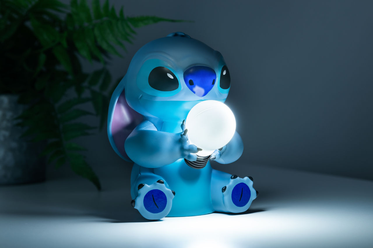Stitch-Lampe, die eine leuchtende Glühbirne hält, blaue Figur auf weißer Fläche, Pflanze im Hintergrund.