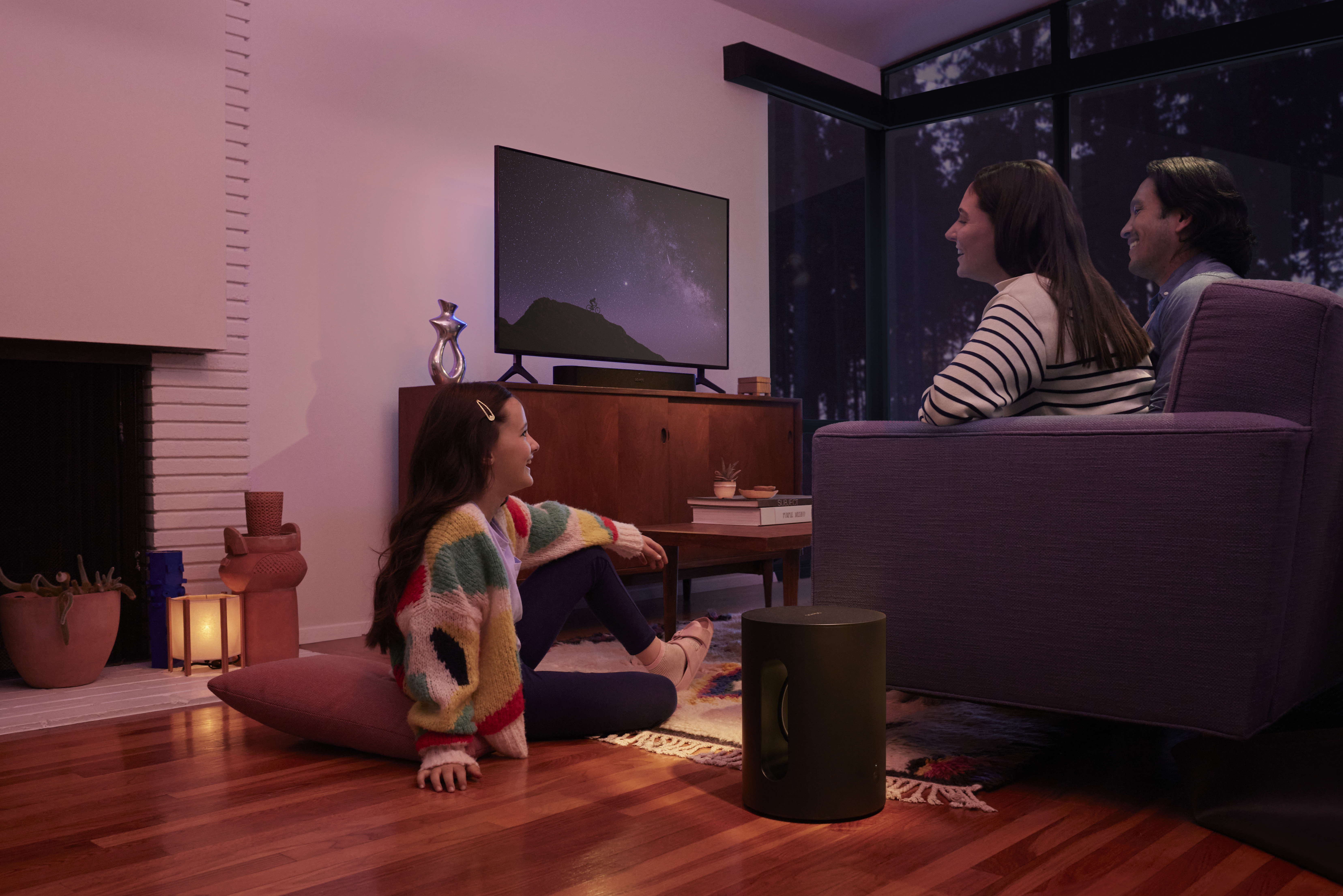 Familie schaut im Wohnzimmer fern. Ein Subwoofer steht auf dem Boden. Es gibt einen Kamin und ein Fenster.