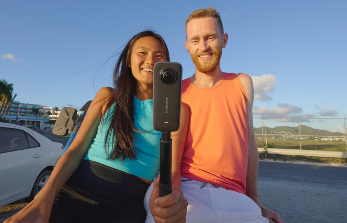 Ein Paar mit einer 360-Grad-Kamera auf einem Selfie-Stick posiert im Freien und lächelt.