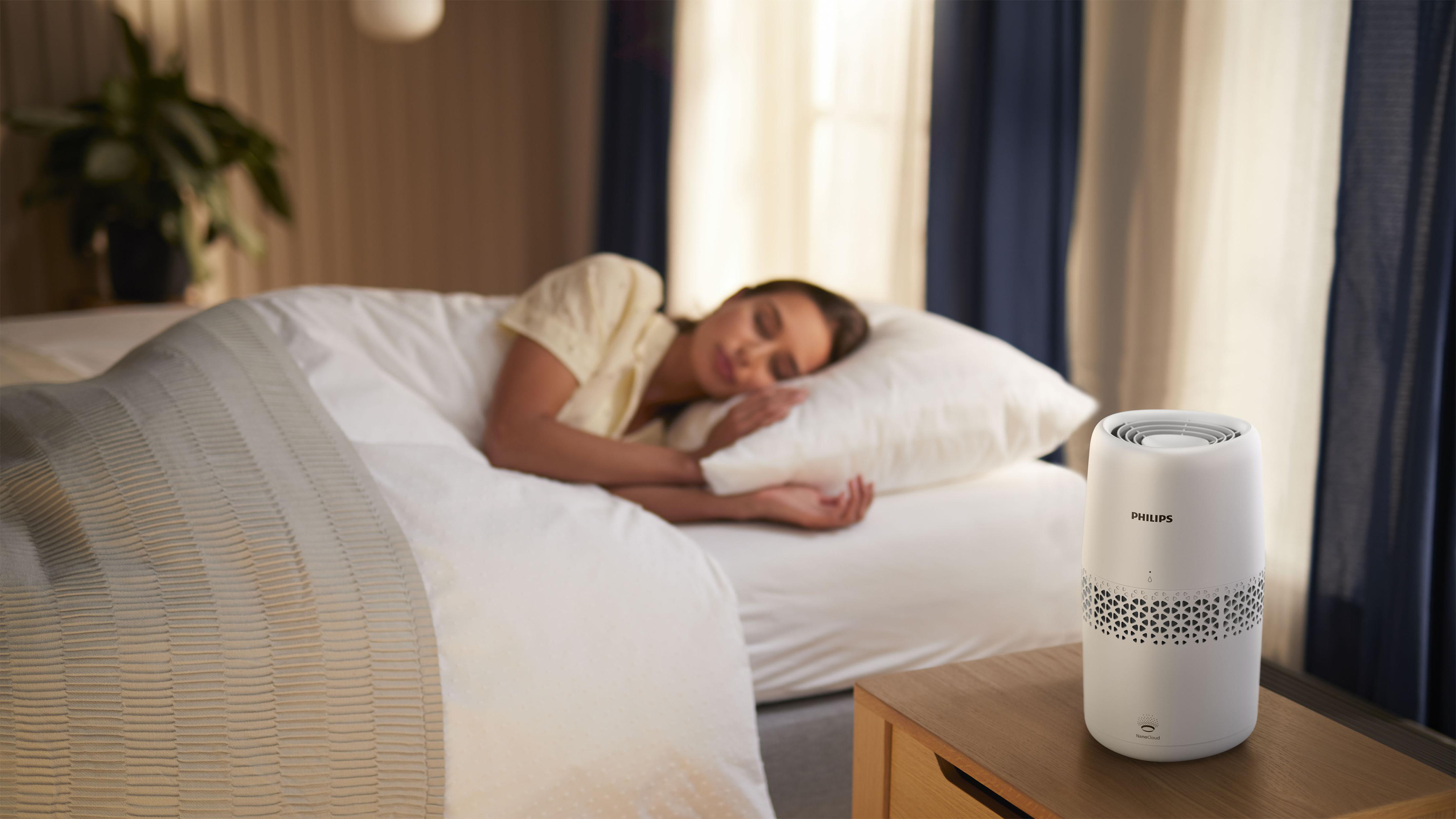 Frau schläft im Bett mit einem weißen Luftreiniger auf einem Nachttisch. Schlafzimmer, tagsüber.