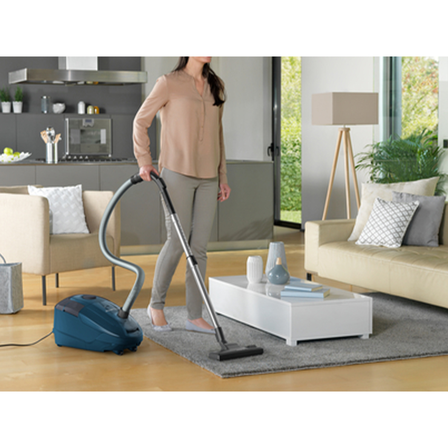 Donna che passa l'aspirapolvere in un soggiorno con un aspirapolvere blu. Divano beige e tavolino bianco visibili.