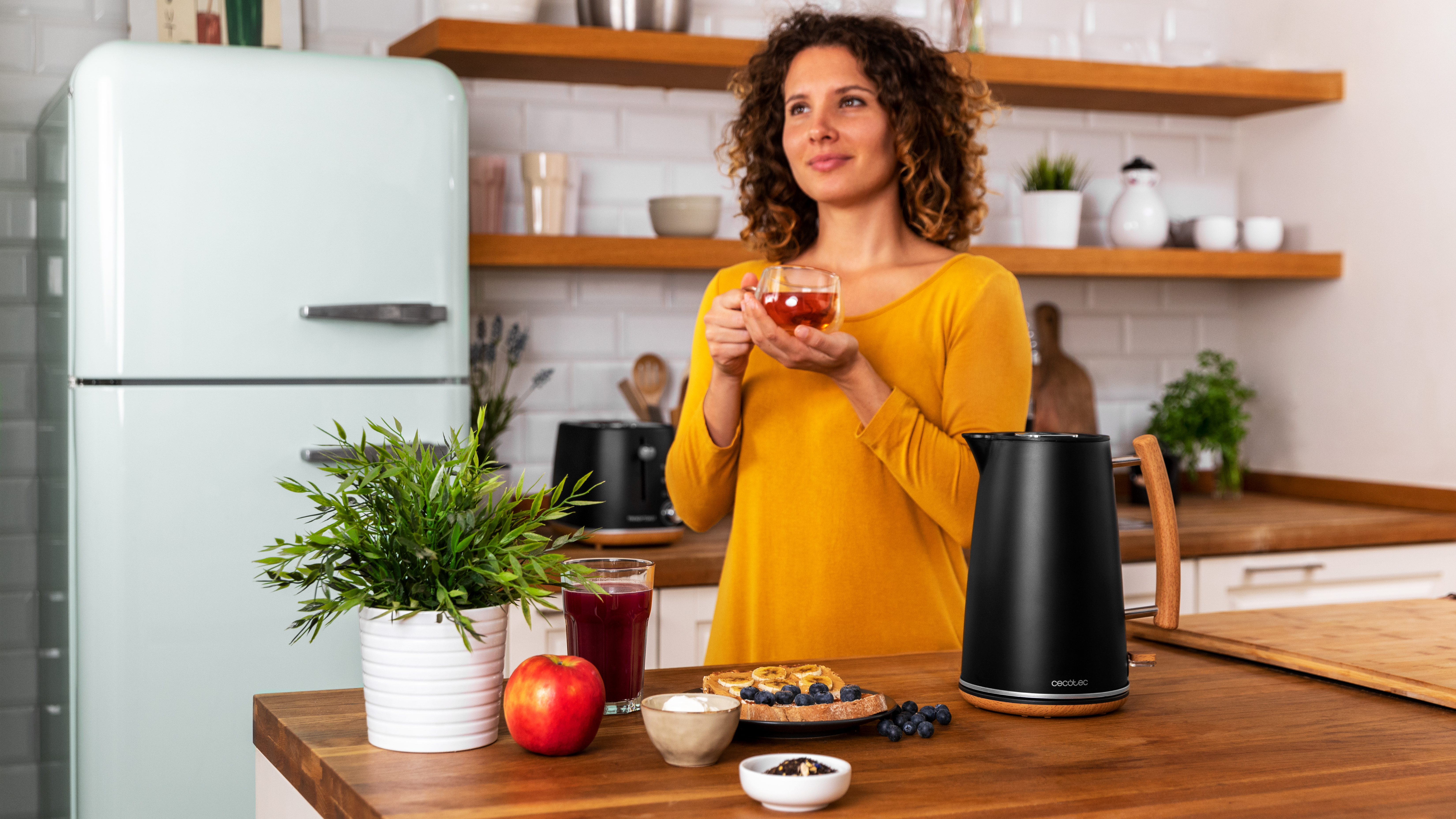 Una mujer con camisa amarilla en una cocina, sosteniendo té, con comida en la mesa.