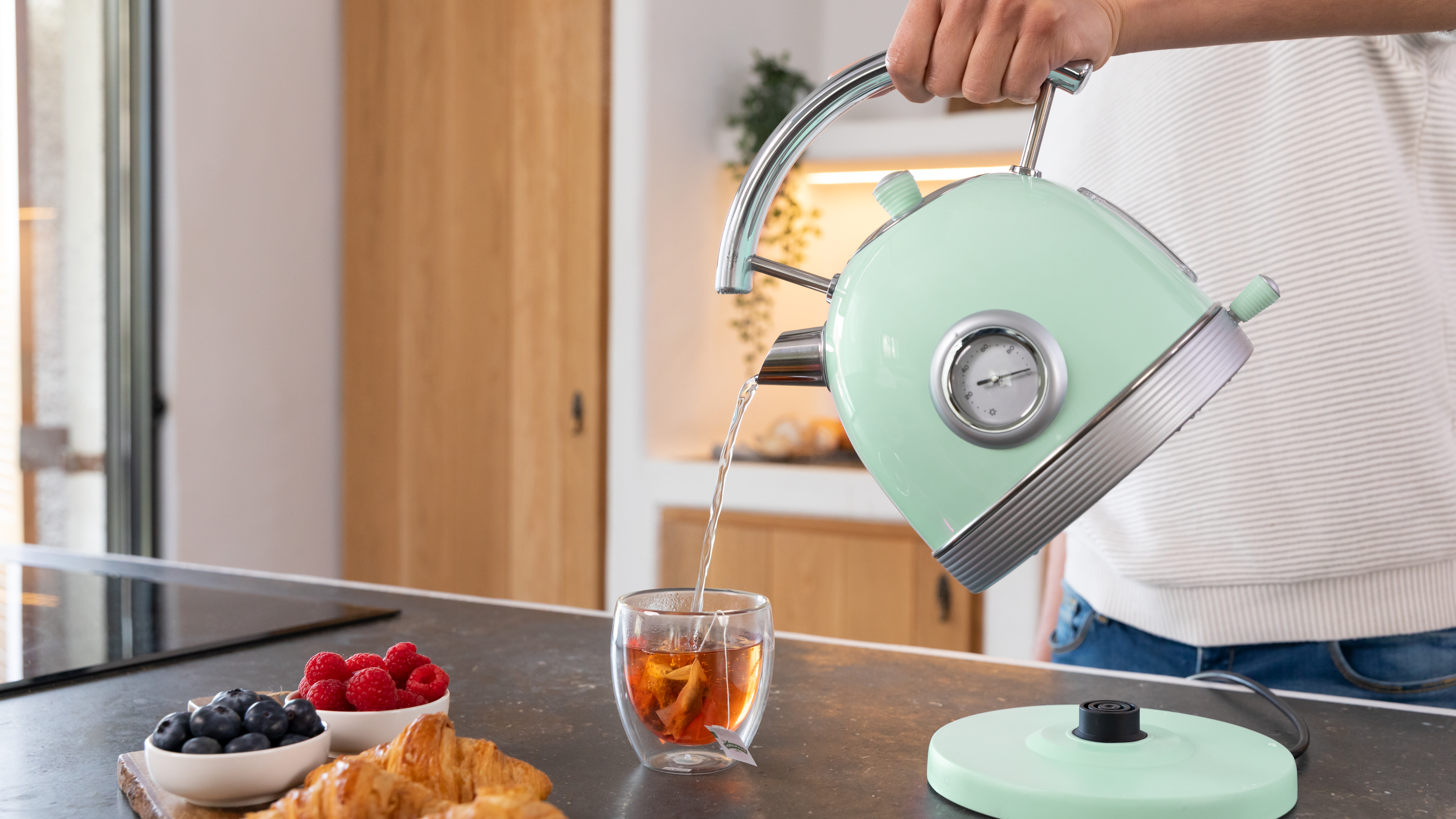 Una persona vierte agua de una tetera verde menta en una taza de té, con pasteles y bayas cerca.