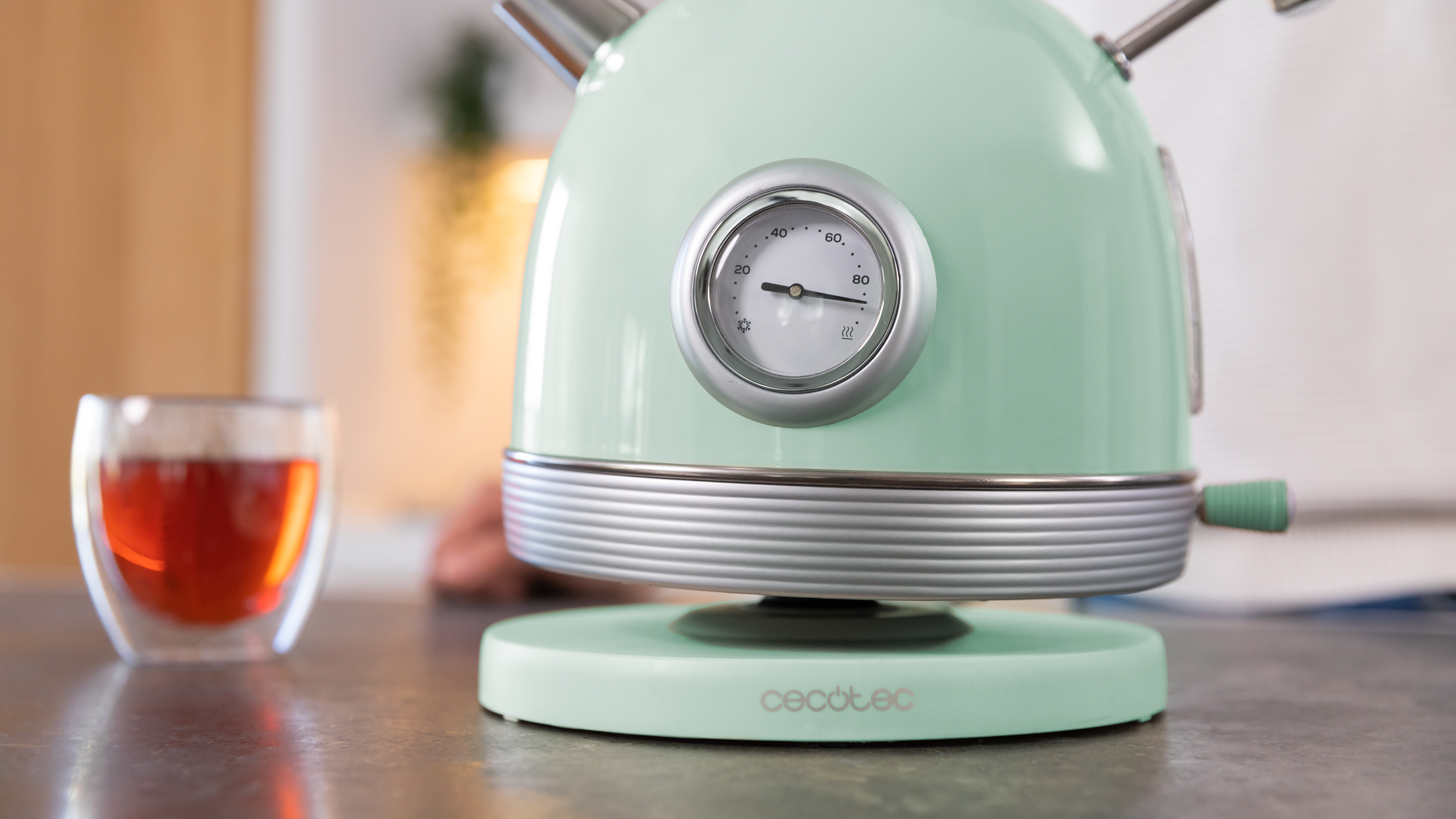 Una tetera verde menta con un indicador de temperatura, colocada sobre una base verde menta. Una taza de vidrio con té al lado.