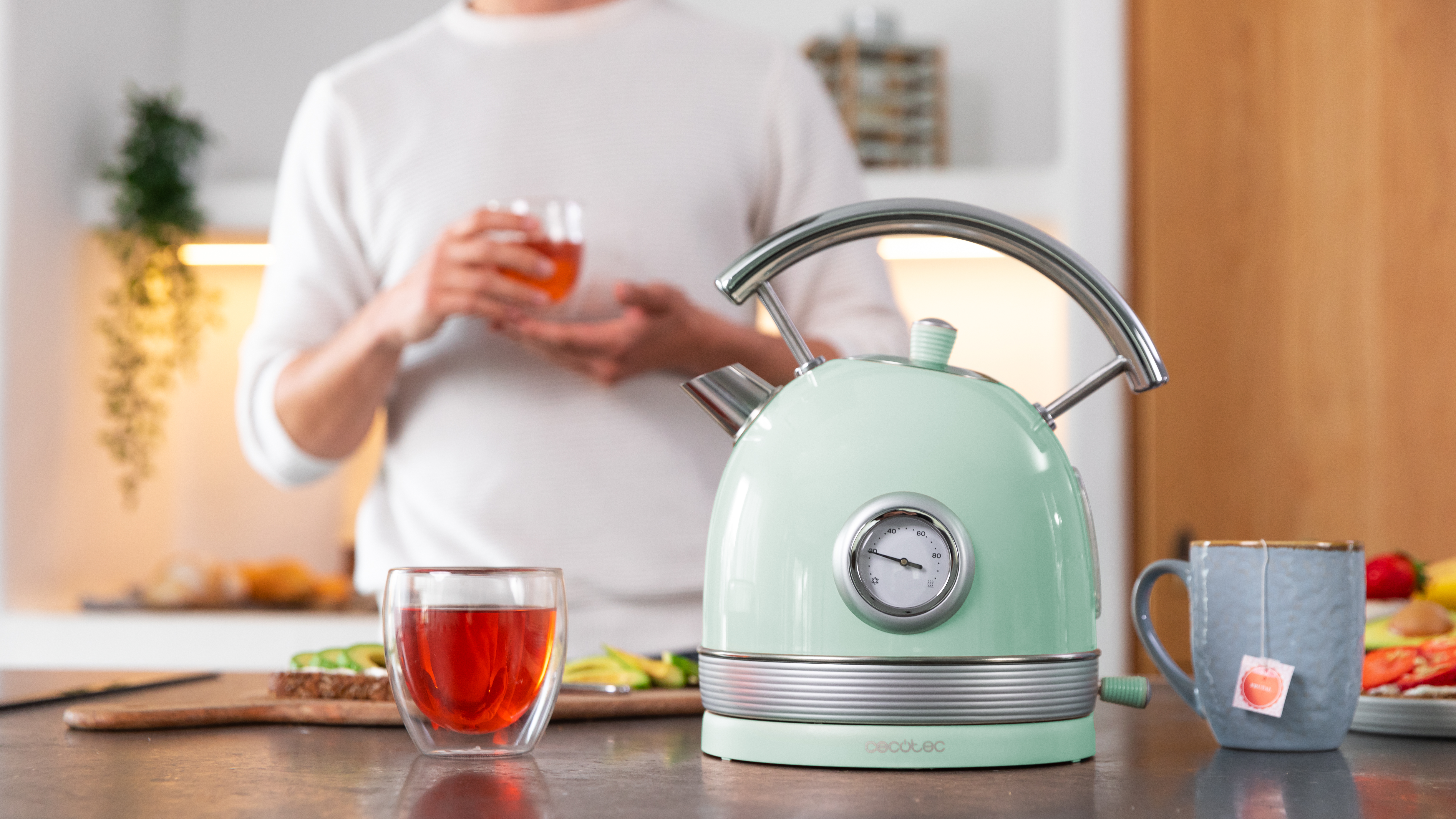 Una persona sosteniendo una taza de té frente a una tetera verde claro y un vaso de té.