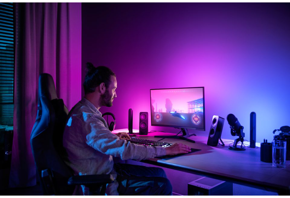 A man gaming at a desk with a monitor, speakers, and microphone in a purple-blue room.