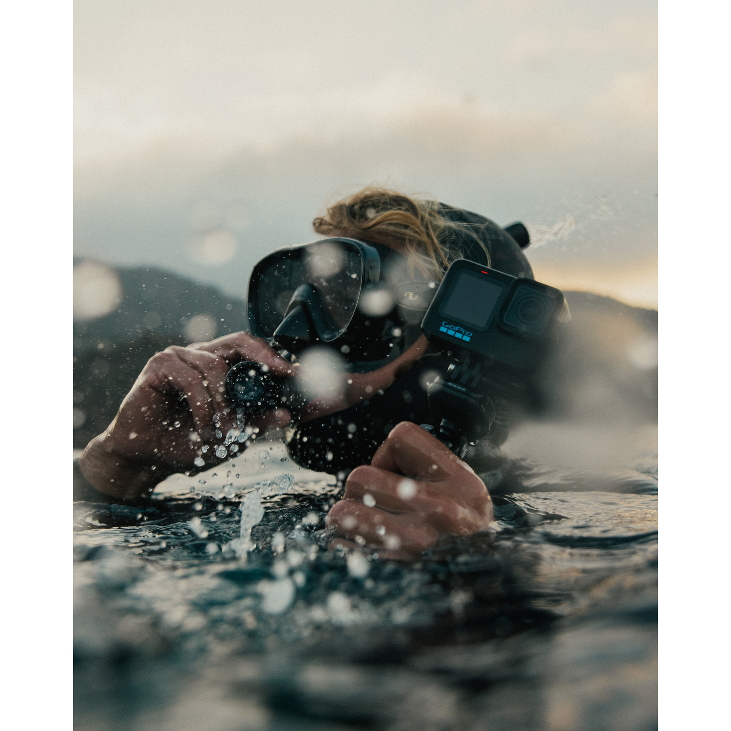 Una persona en el agua lleva una máscara de buceo y una GoPro. El agua salpica a su alrededor. Cielo nublado al fondo.