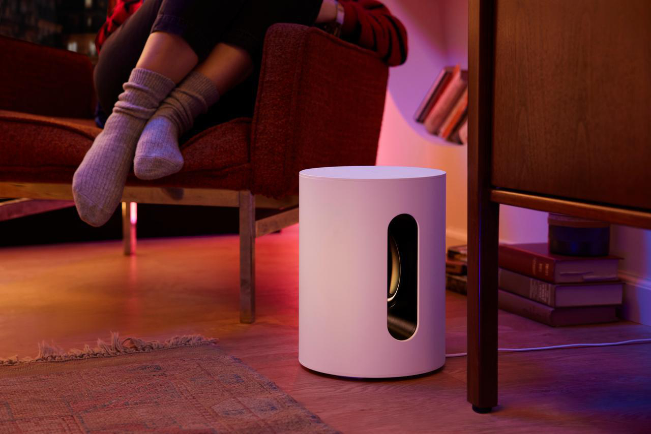 Weißer zylindrischer Subwoofer mit schwarzer Öffnung. Frau sitzt im Hintergrund.