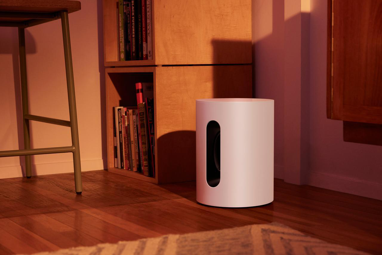 Ein weißer Subwoofer-Lautsprecher mit einer Öffnung, neben einem Bücherregal und einer Holzoberfläche.