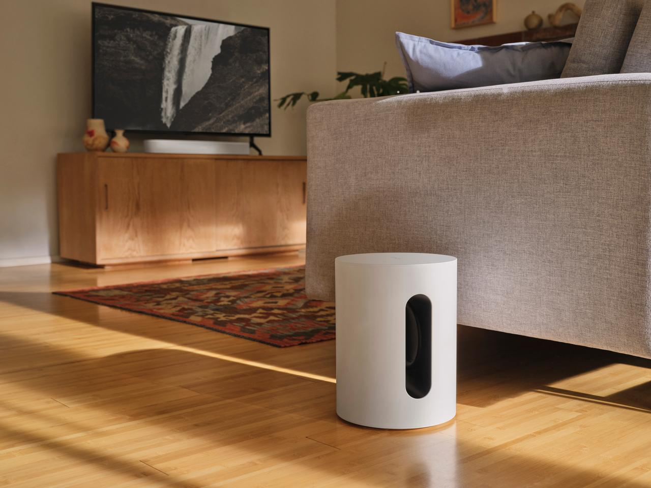 Ein weißer Subwoofer-Lautsprecher neben einem Sofa und einem Fernseher in einem Wohnzimmer.
