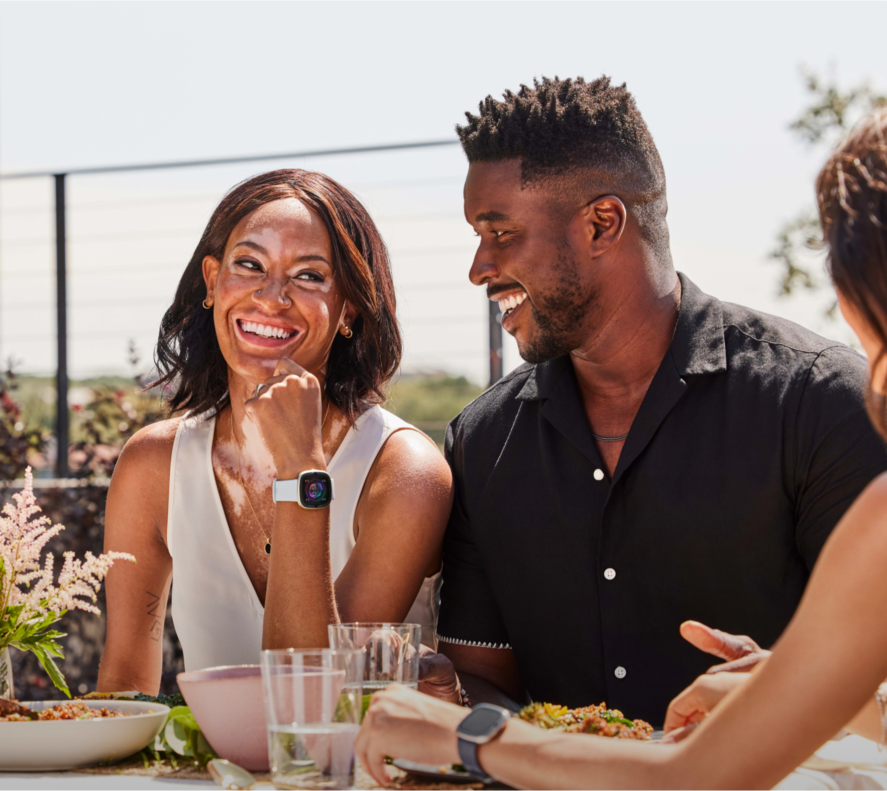 A mixed-race group enjoys a meal outdoors. Two people are laughing. One wears a watch. Food and drinks are on the table.