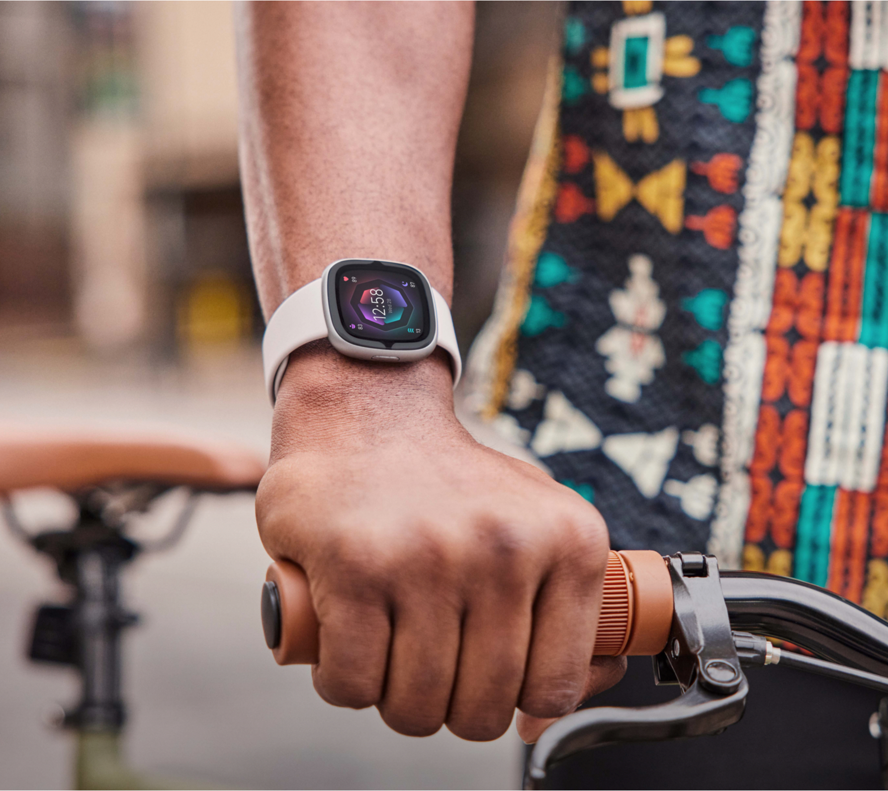 A person's arm holding a bike handlebar, wearing a smartwatch. The watch displays time. Background has buildings and a patterned shirt.