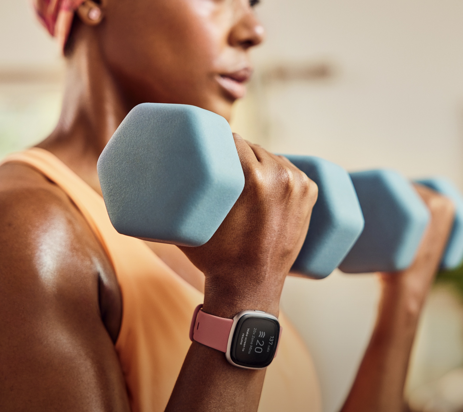 A woman lifts blue dumbbells. She wears a pink watch and orange top. Background is slightly blurred.