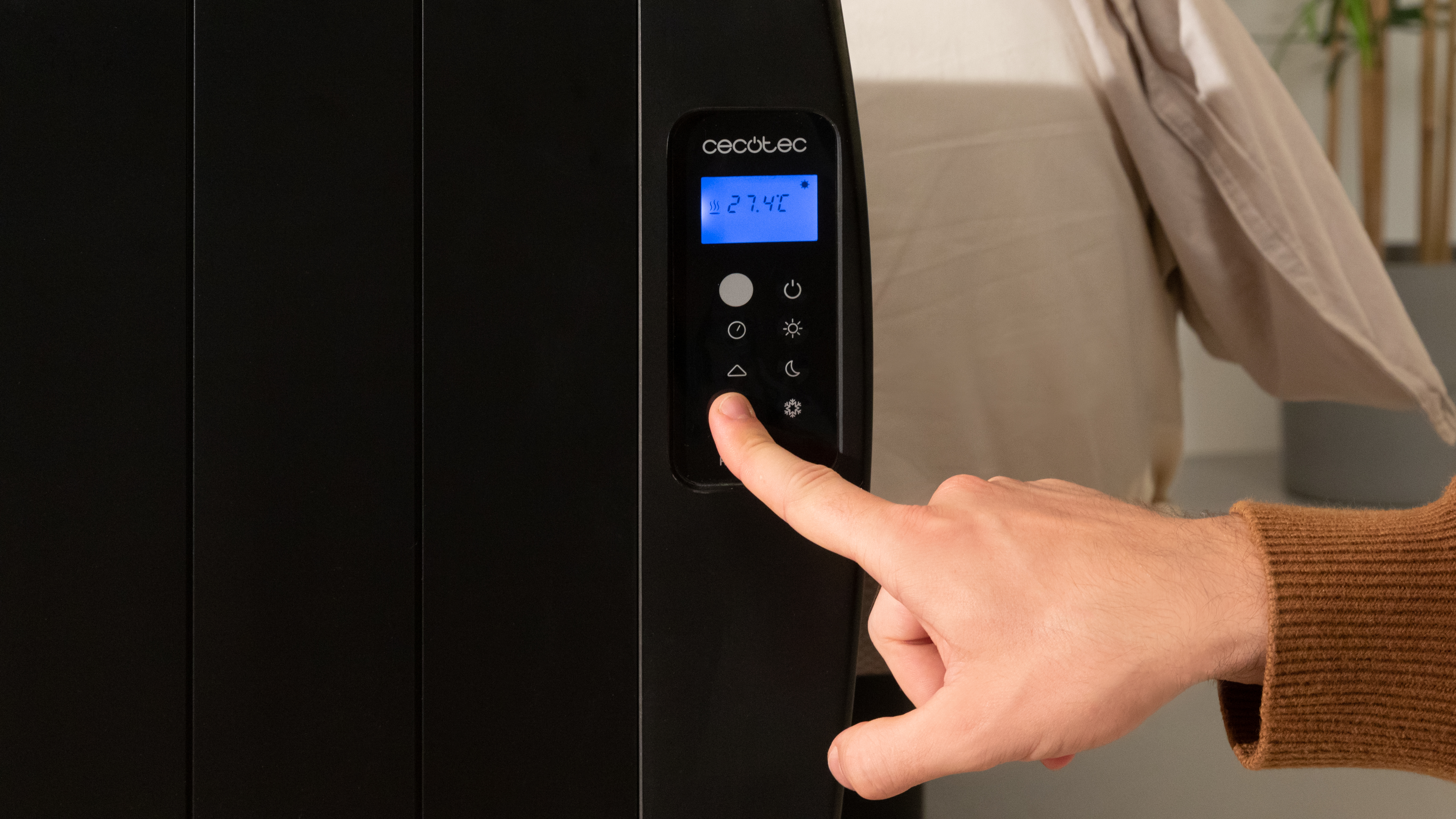 La mano de una persona toca el panel de control de un radiador negro. La pantalla muestra la temperatura.