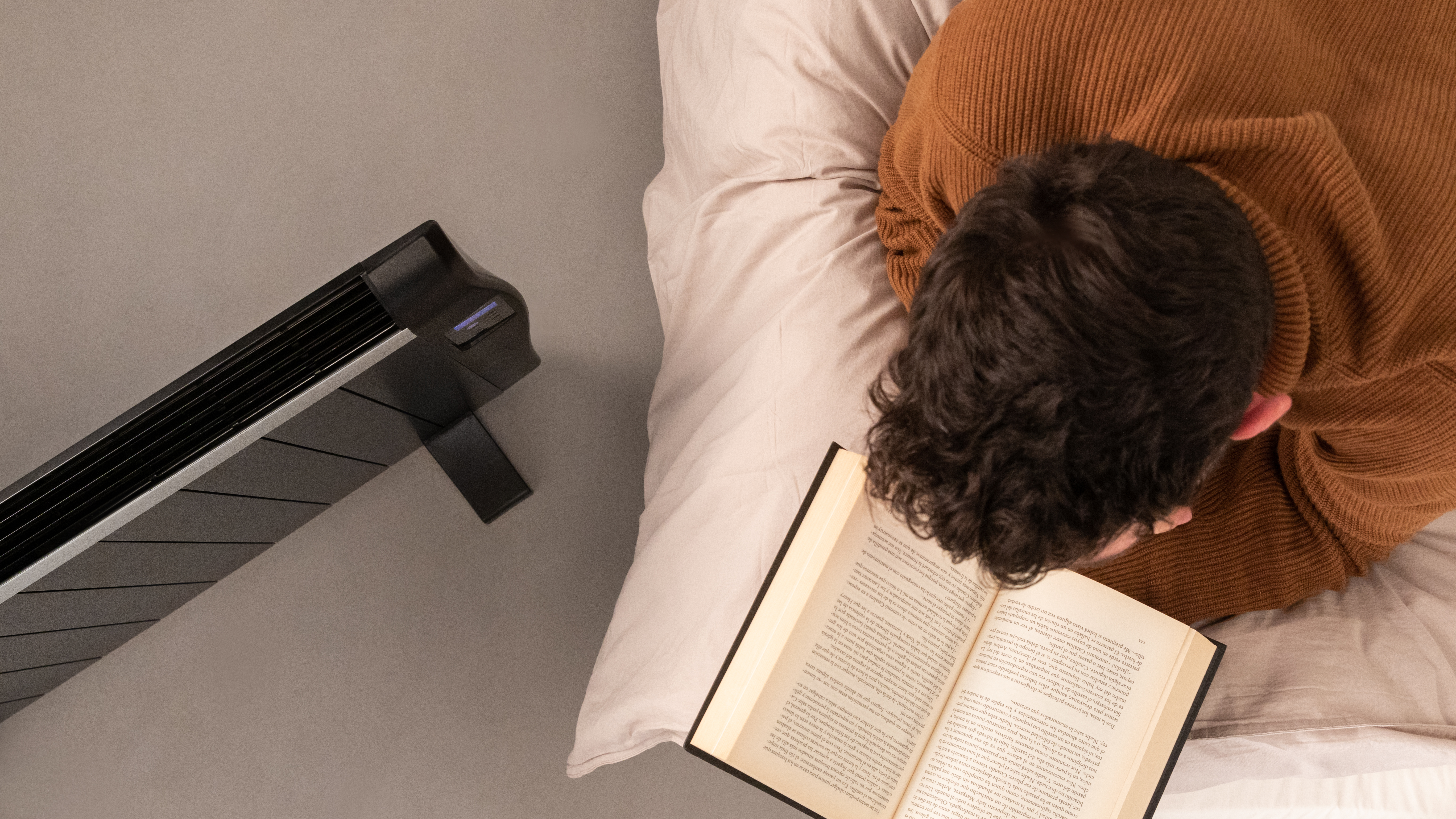 Una persona acostada en la cama con un libro y un calentador al lado; vista superior.