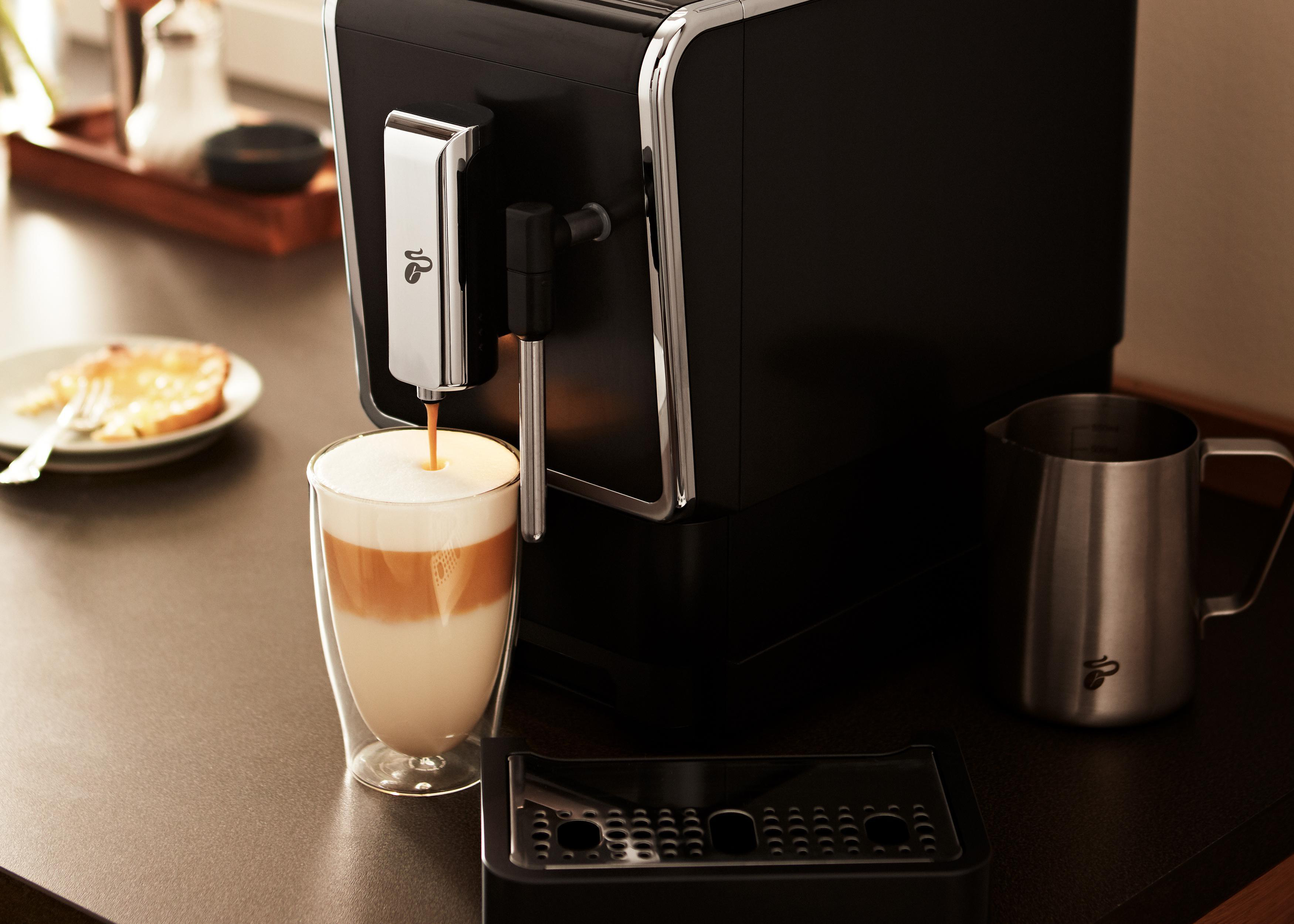 Eine schwarze Kaffeemaschine gießt Kaffee in ein Glas. Links ist ein Milchaufschäumer.