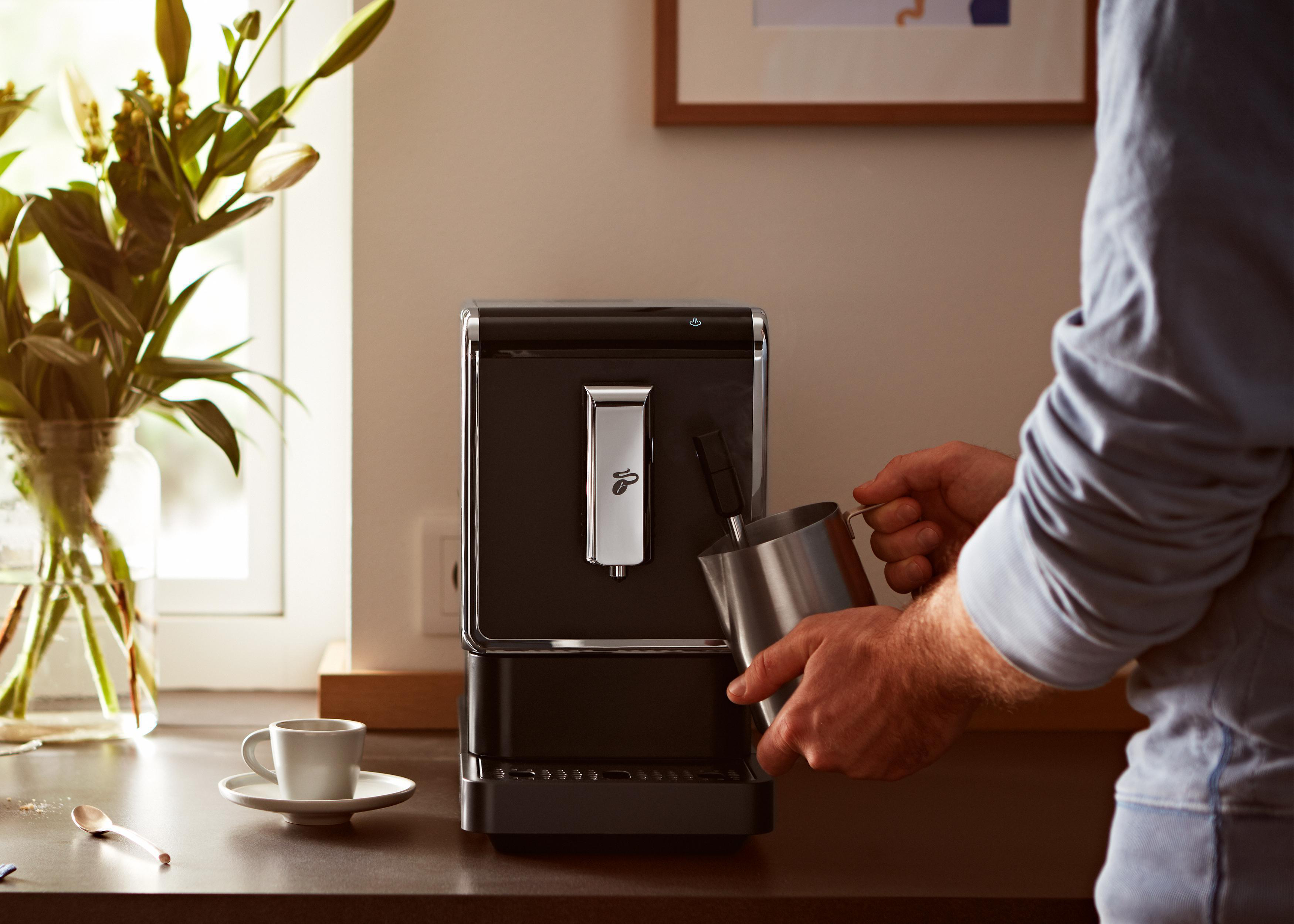 Eine Person gießt Milch in eine Kaffeemaschine. Eine Tasse und ein Löffel stehen auf der Theke.