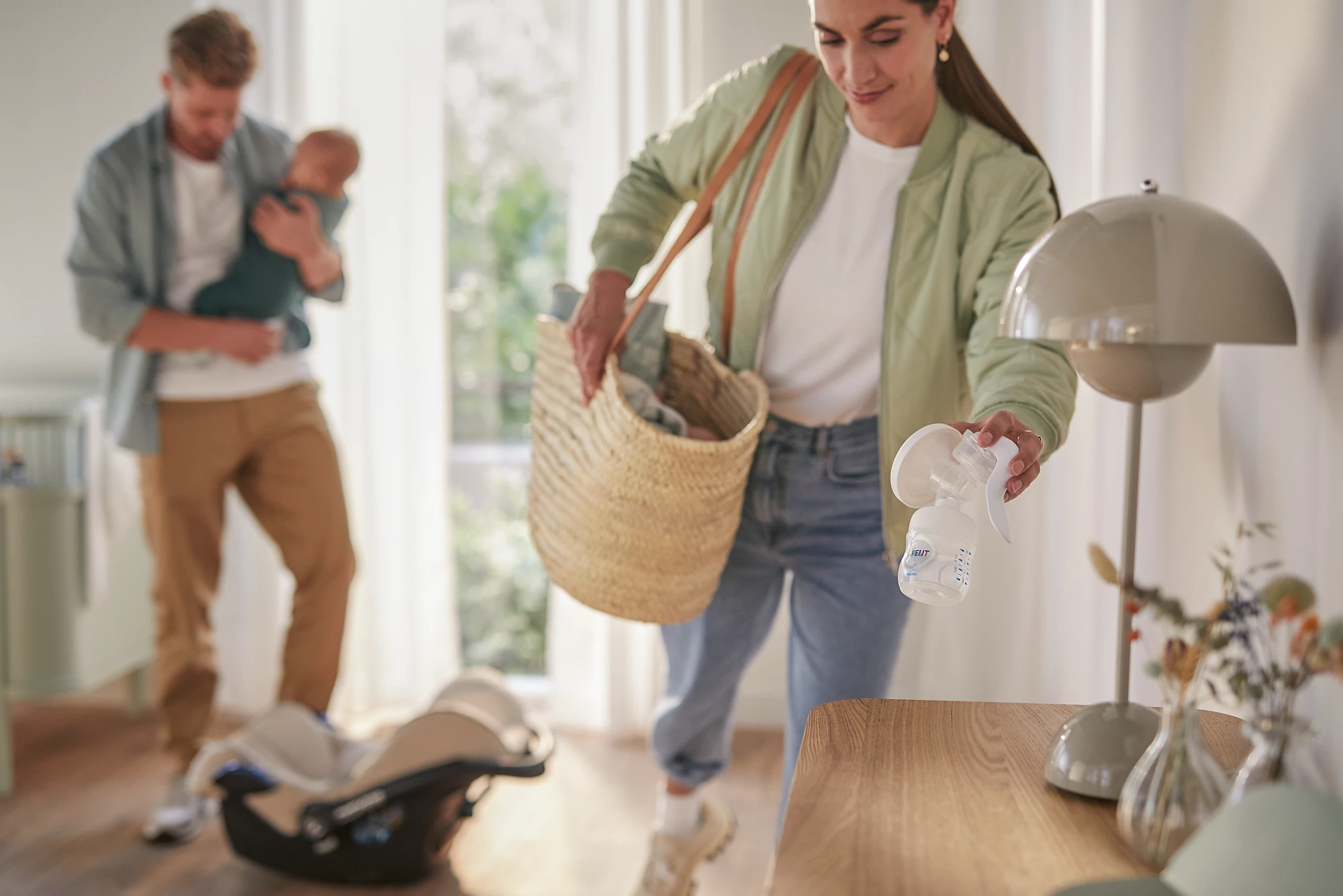 Frau mit Baby und ein Mann im Hintergrund. Eine Lampe steht auf einem Tisch.
