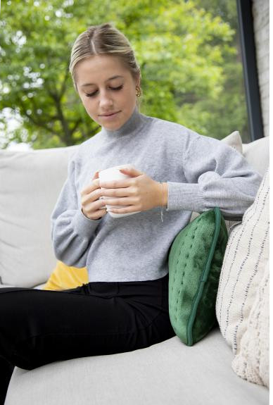 Femme tenant une tasse blanche, assise sur un canapé. Elle porte un pull gris et un pantalon noir.