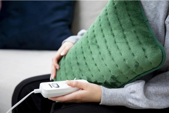 Une femme tient un coussin chauffant vert avec un panneau de commande dans ses mains, assise sur un canapé.