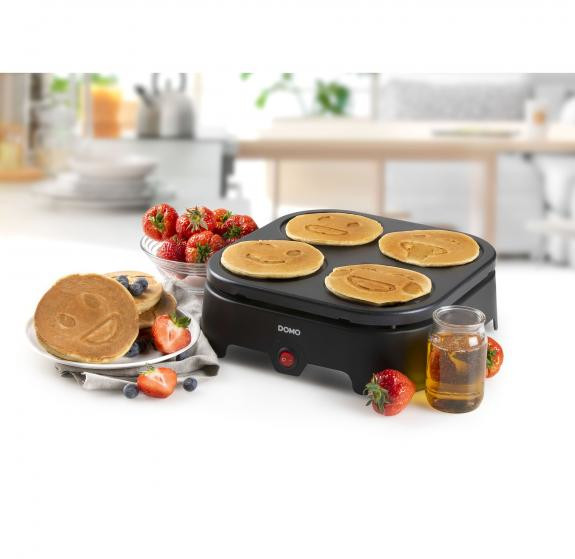 Une machine à crêpes avec des crêpes à smiley. Fraises, sirop et myrtilles sont sur le côté.
