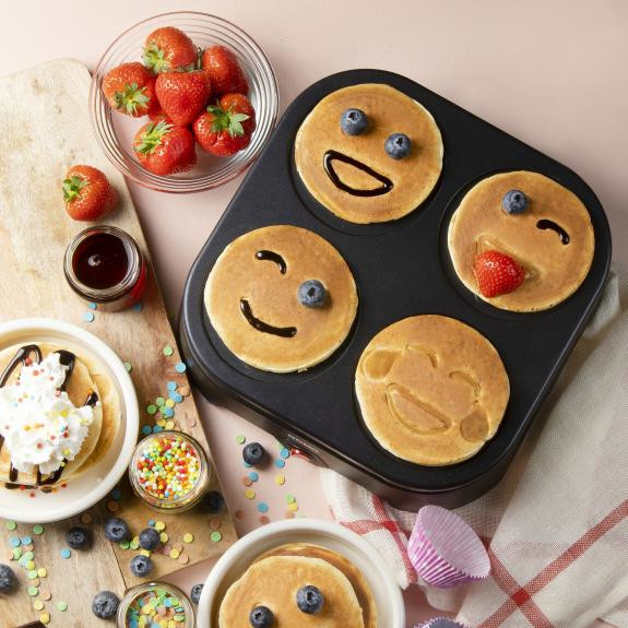 Machine à pancakes avec pancakes souriants, fraises, myrtilles et garnitures sur une surface en bois.