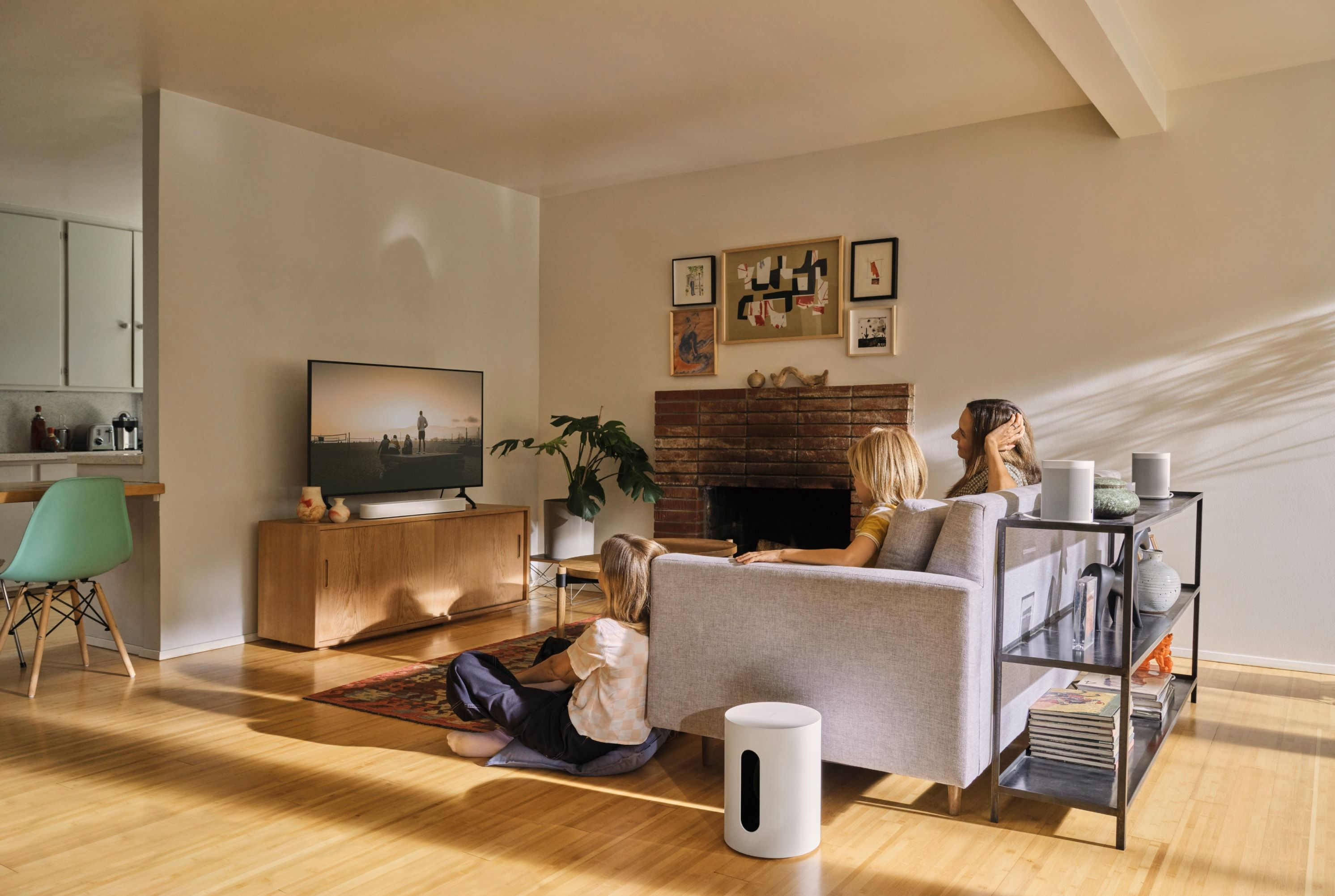 Persone sedute su un divano e per terra che guardano la TV. Un subwoofer bianco e una mensola nera sono nella stanza.