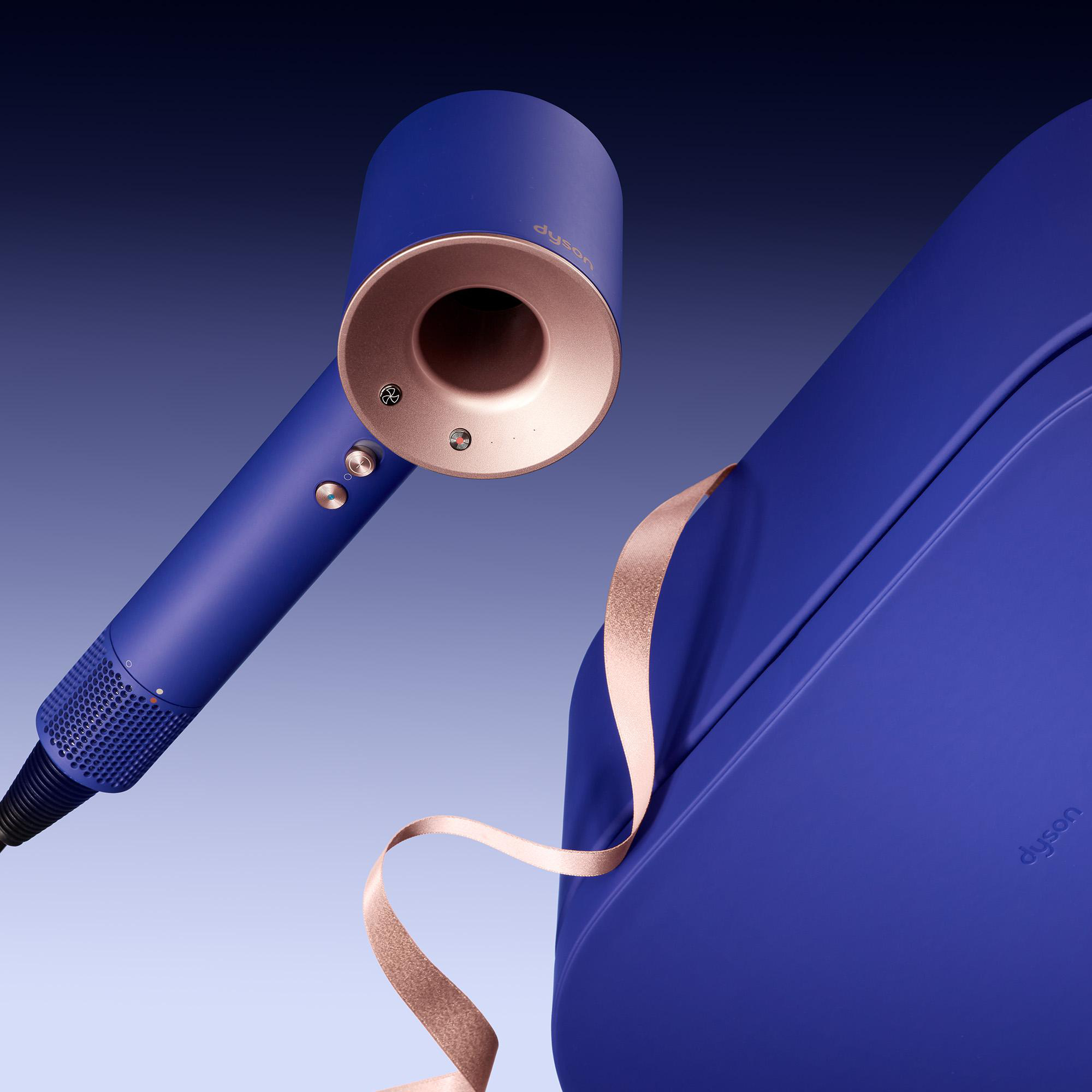 Ein blauer Dyson-Haartrockner mit Kupferdüse neben einem blauen Koffer mit rosa Band.