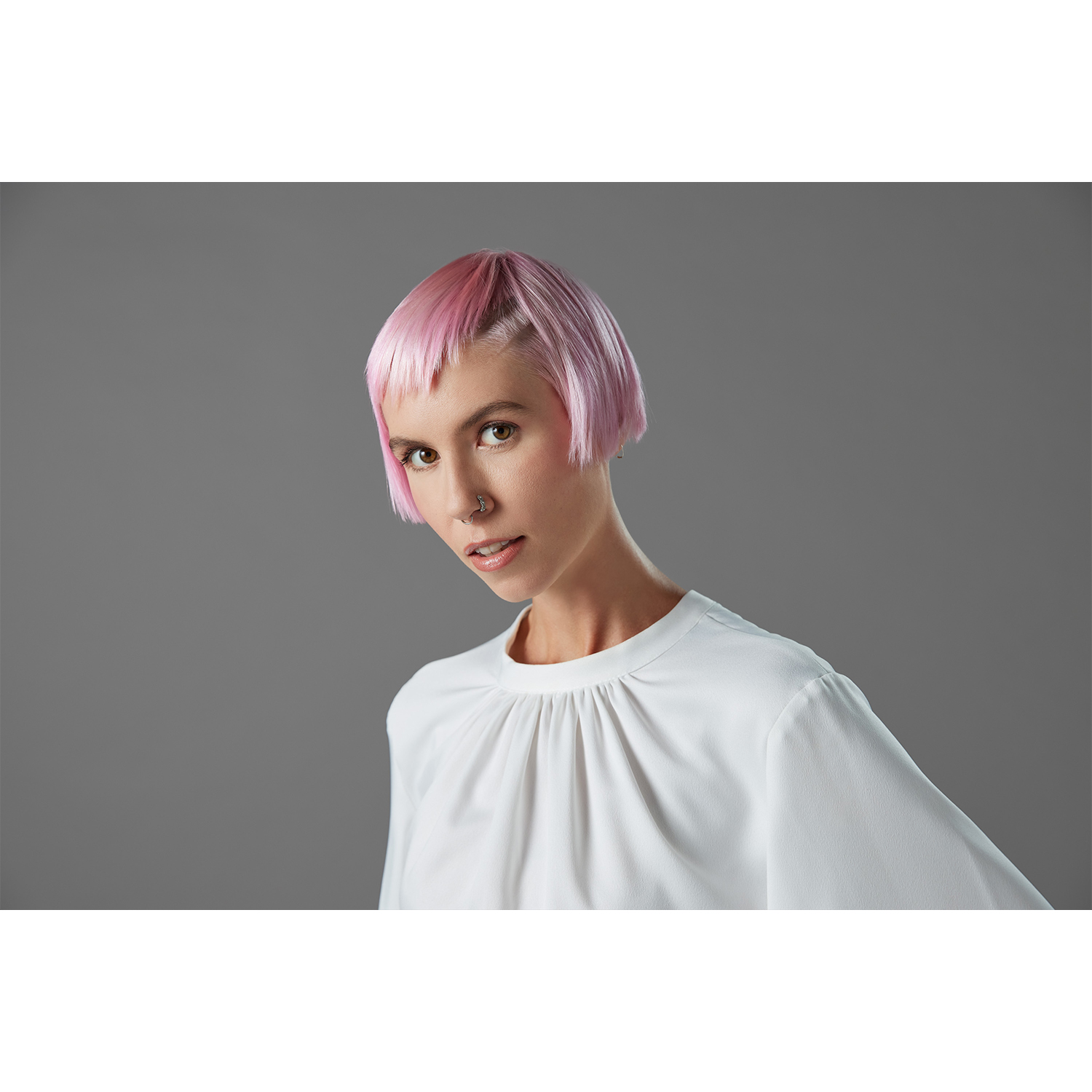 Una donna con i capelli rosa e un piercing al naso guarda lo spettatore. Indossa una camicetta bianca.
