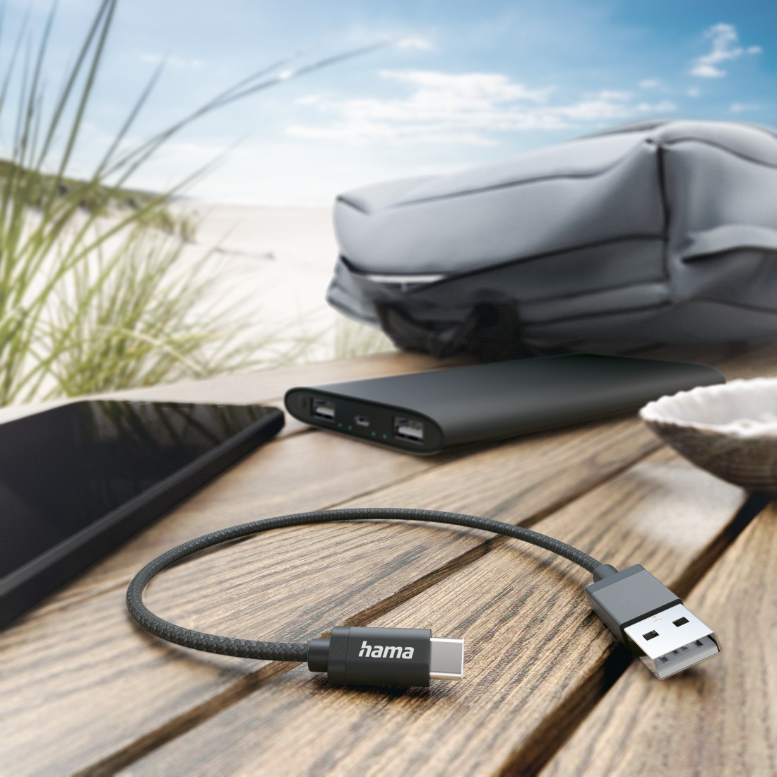 USB kábel, power bank és kiegészítők egy fa asztalon, a háttérben a tengerparttal.