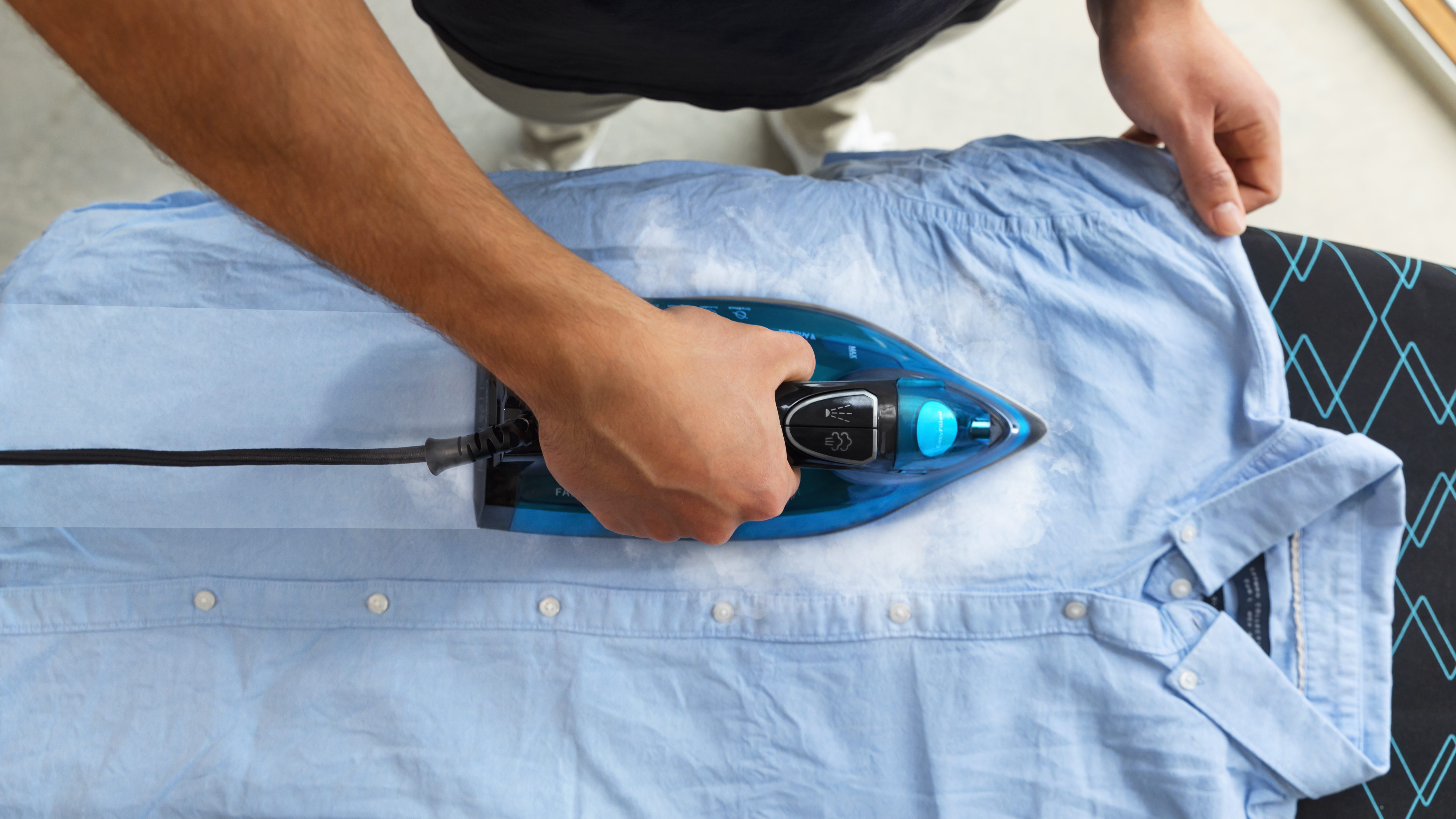 Una persona plancha una camisa azul claro sobre una tabla de planchar. La plancha es azul y emite vapor. La tabla tiene un patrón negro y azul.