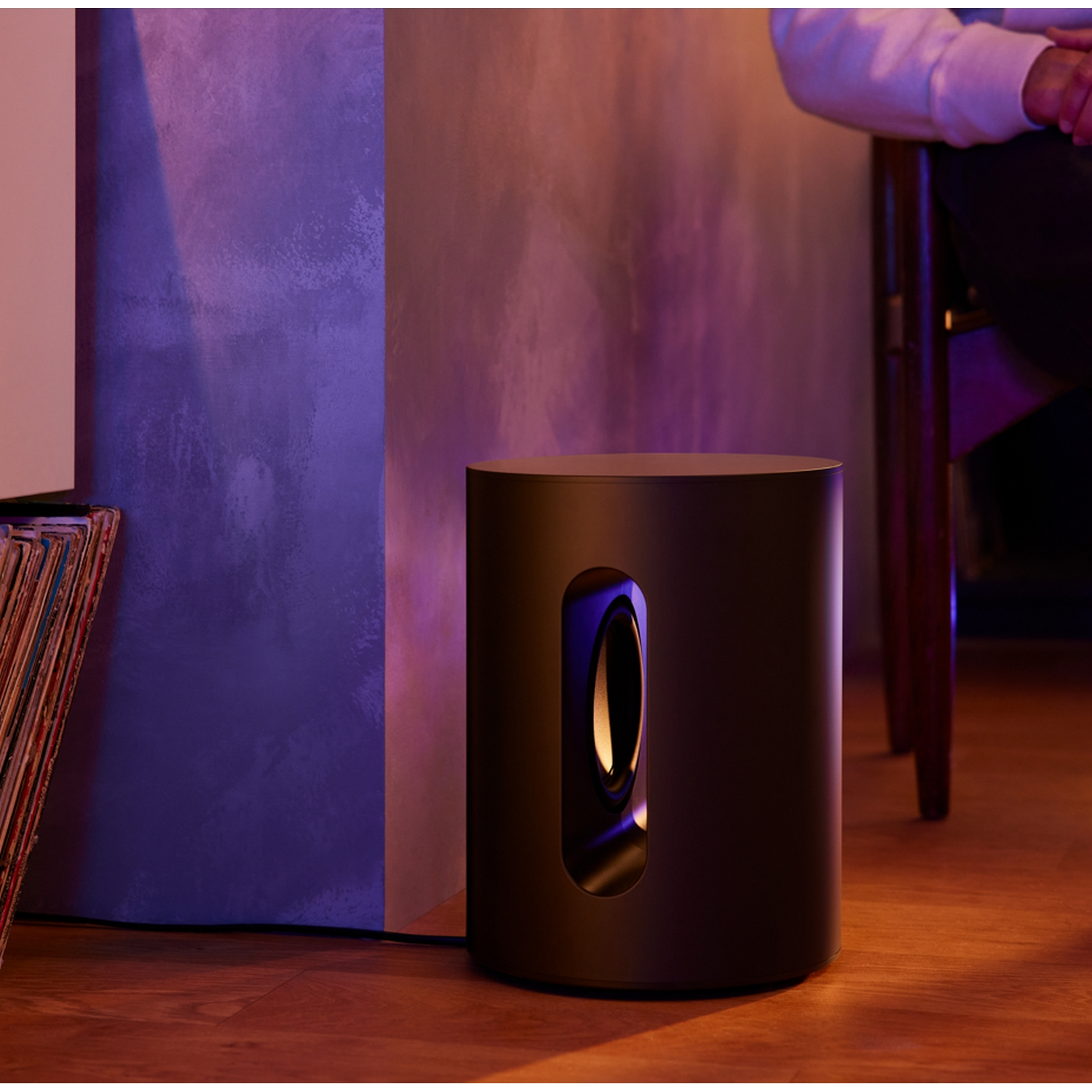 Subwoofer cilíndrico negro con abertura ovalada central, en interiores.