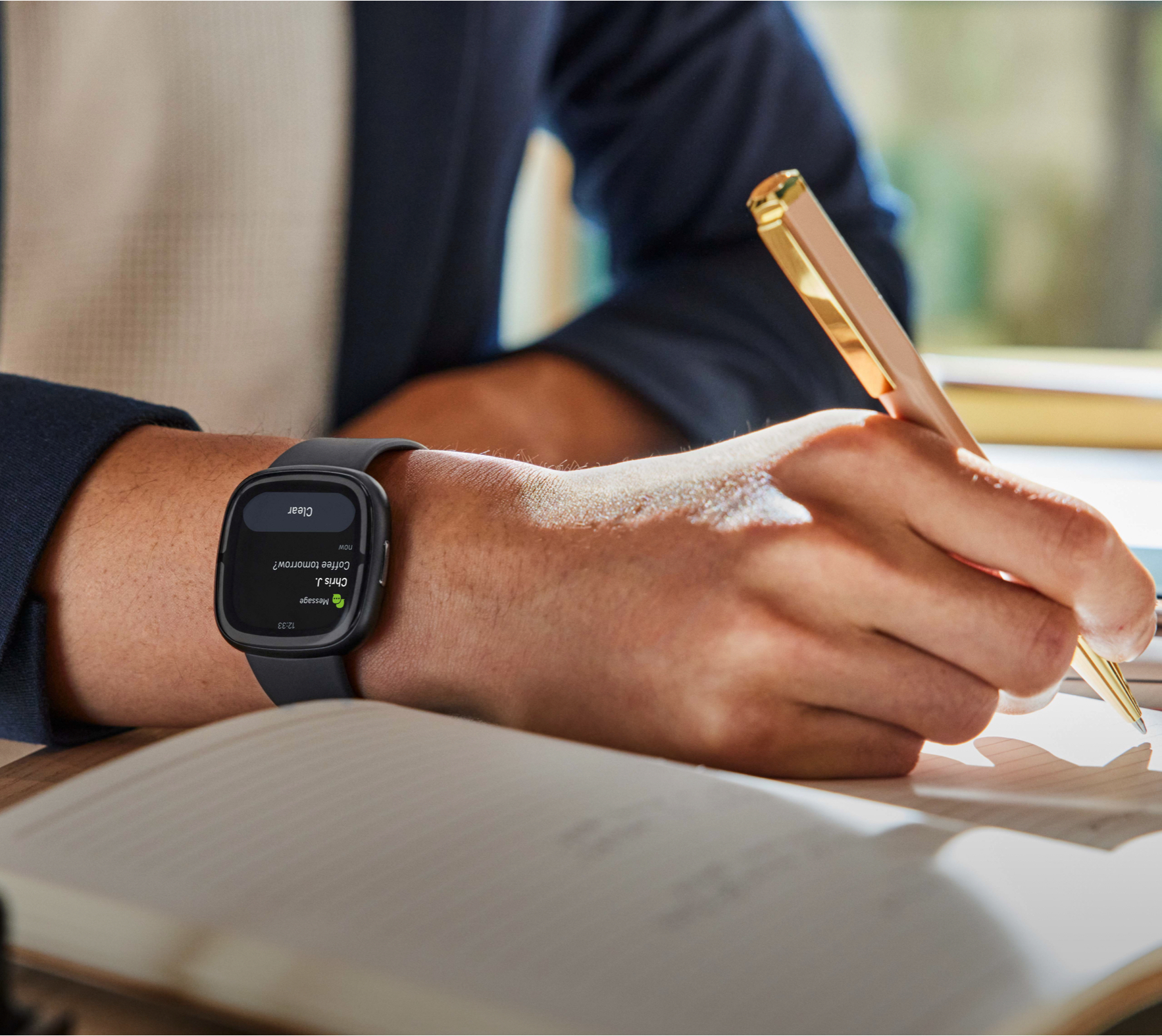 Personne écrit dans un cahier avec un stylo, portant une smartwatch. La montre affiche infos.