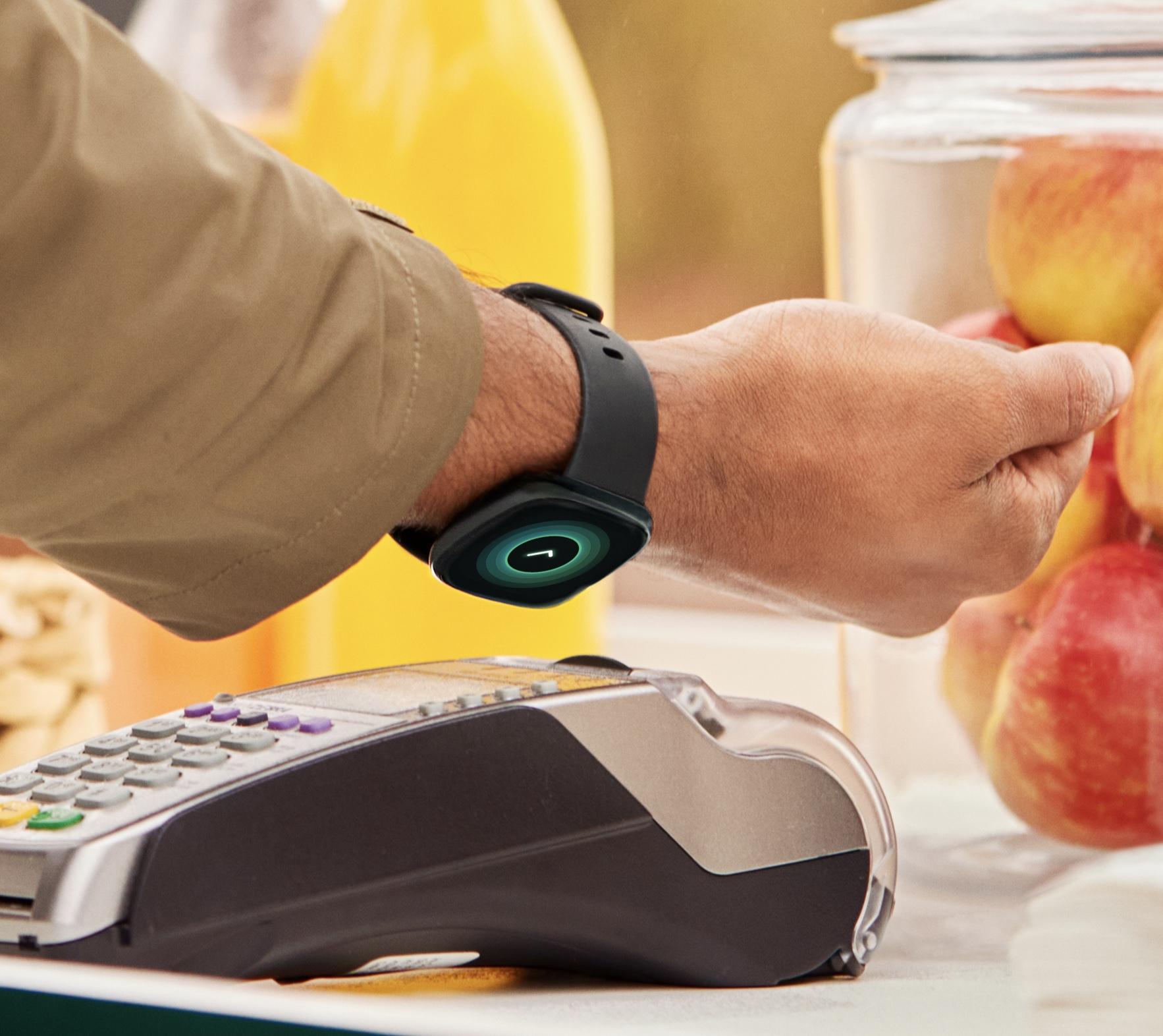 A person is paying with a smartwatch. A payment terminal is in the foreground. Apples and juice are in the background.