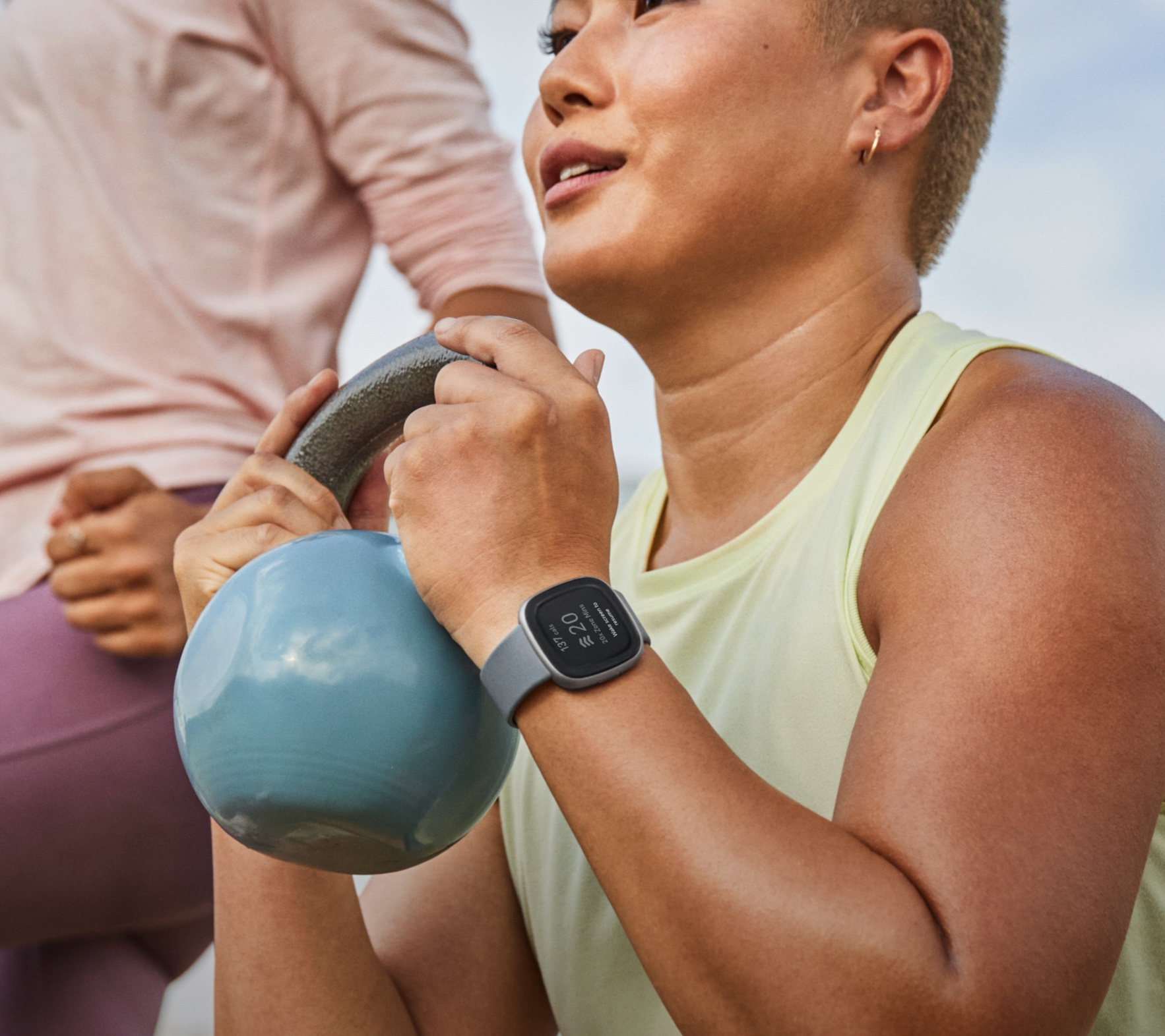 Woman holding a blue kettlebell, wearing a smartwatch. Another person is in the background. Sky visible.