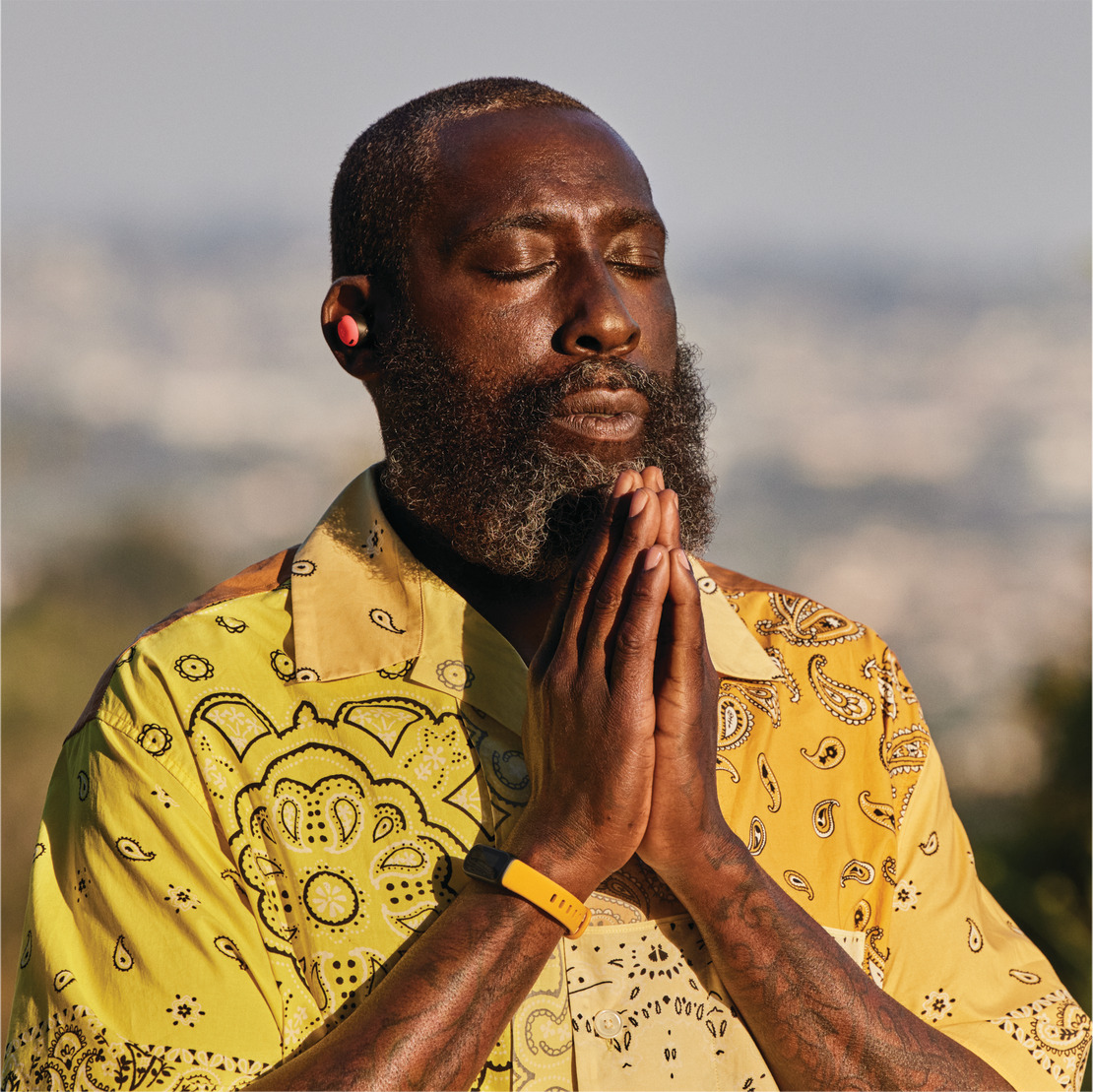 A man with closed eyes and clasped hands is wearing a yellow shirt and orange earbuds, with a blurred background.
