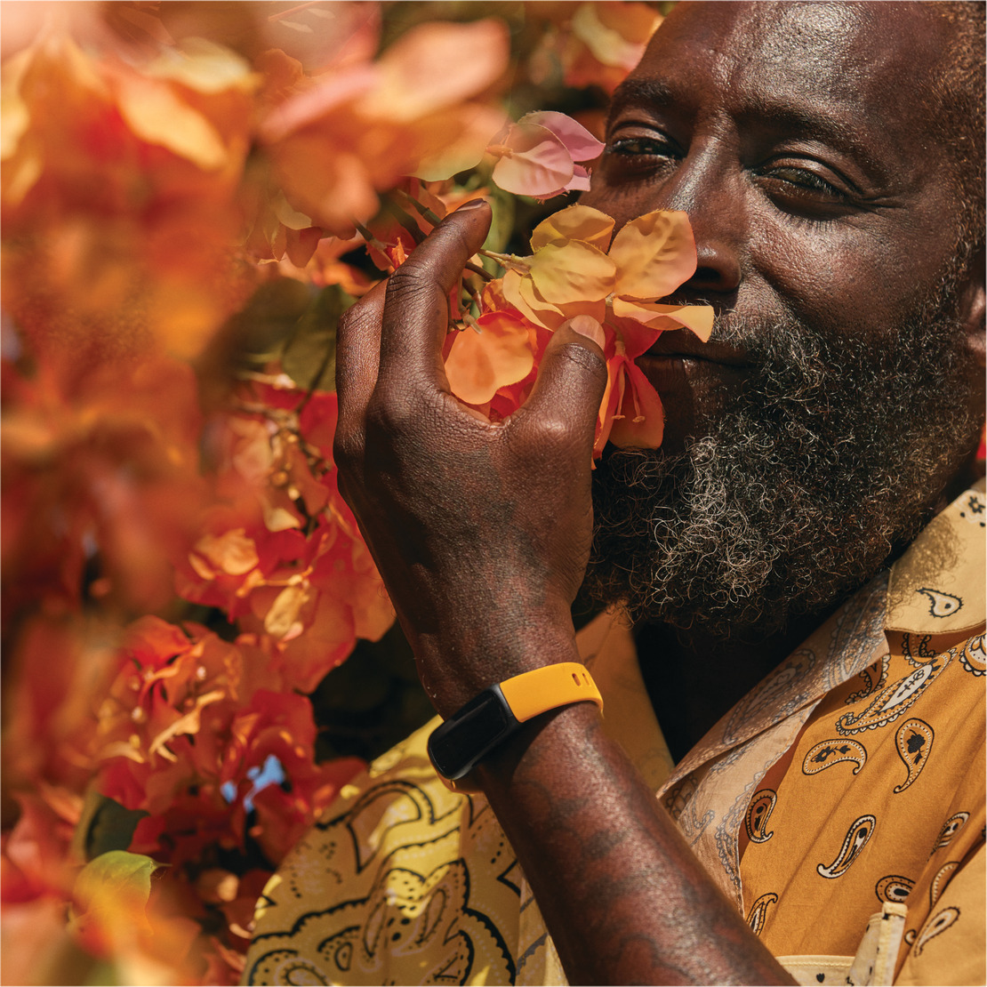 A man smelling orange flowers, wearing a yellow patterned shirt and a black and yellow wristband.
