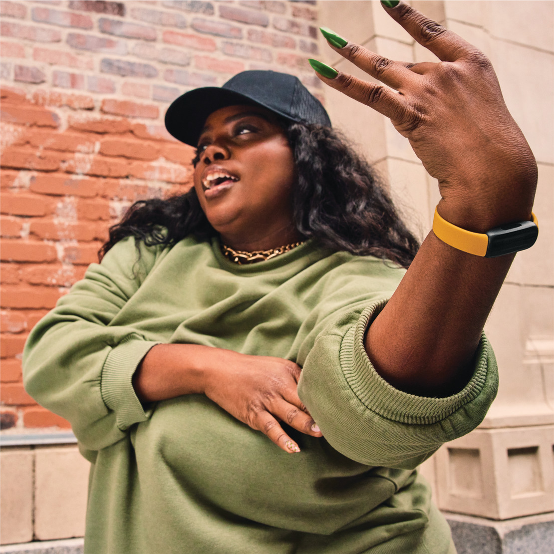 A woman in green sweatshirt and black cap poses. She gestures with a gold and black bracelet.