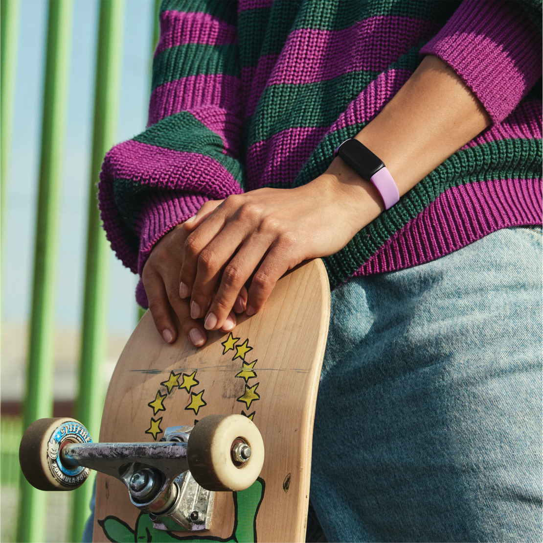 Person holding a skateboard, wearing a purple and green striped sweater and a smartwatch.