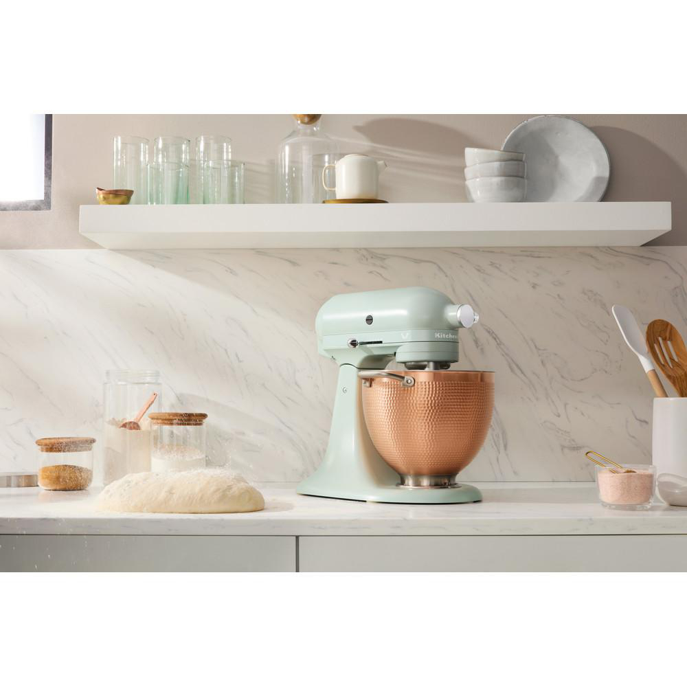 Ein mintgrüner KitchenAid-Küchenmixer mit einer Kupferschüssel, Mehl und Teig auf einer weißen Theke.