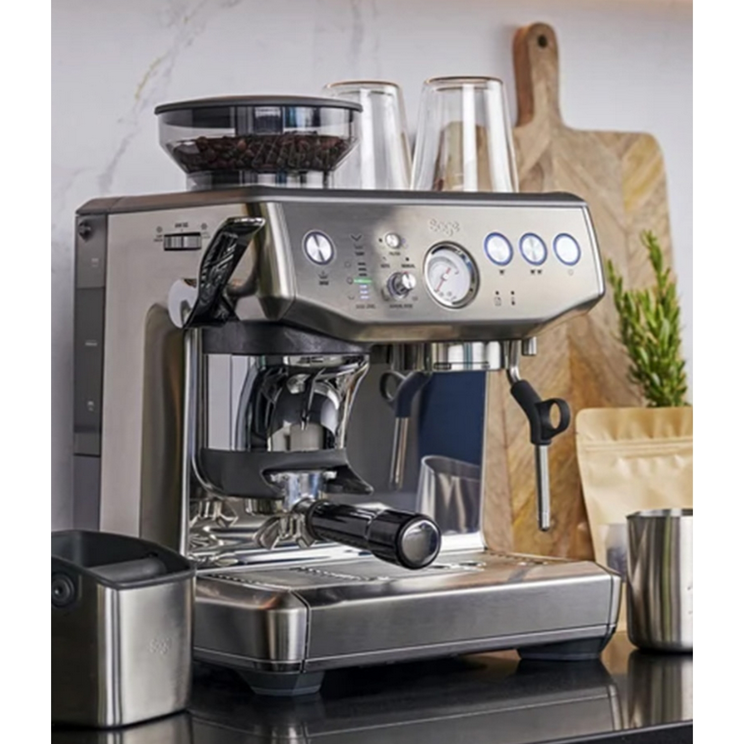 Cafetera espresso plateada con accesorios sobre encimera negra. Tabla de cortar de madera a la derecha.