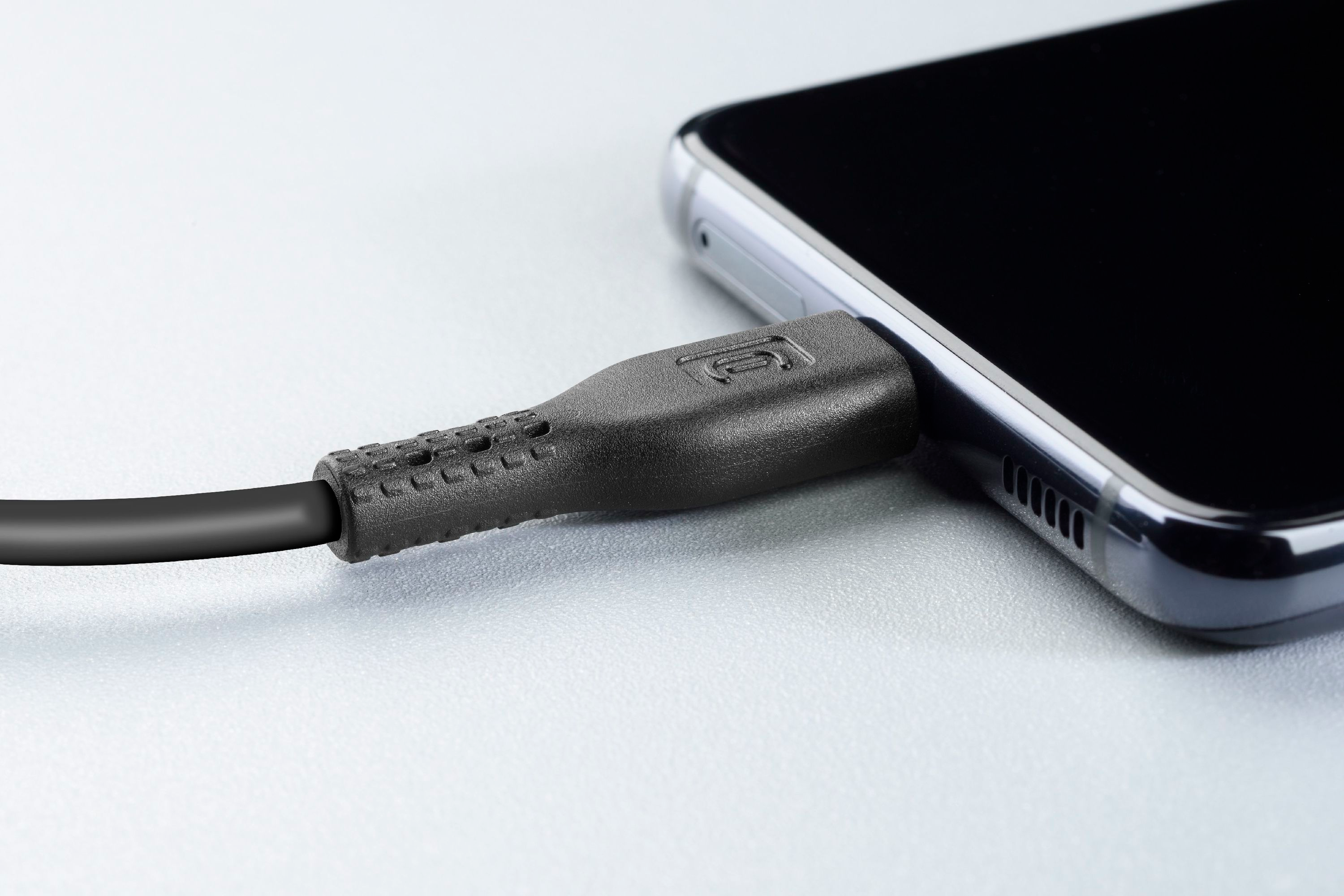 Ein schwarzes Ladekabel, das in ein schwarzes Smartphone eingesteckt ist, auf heller, grauer Oberfläche.