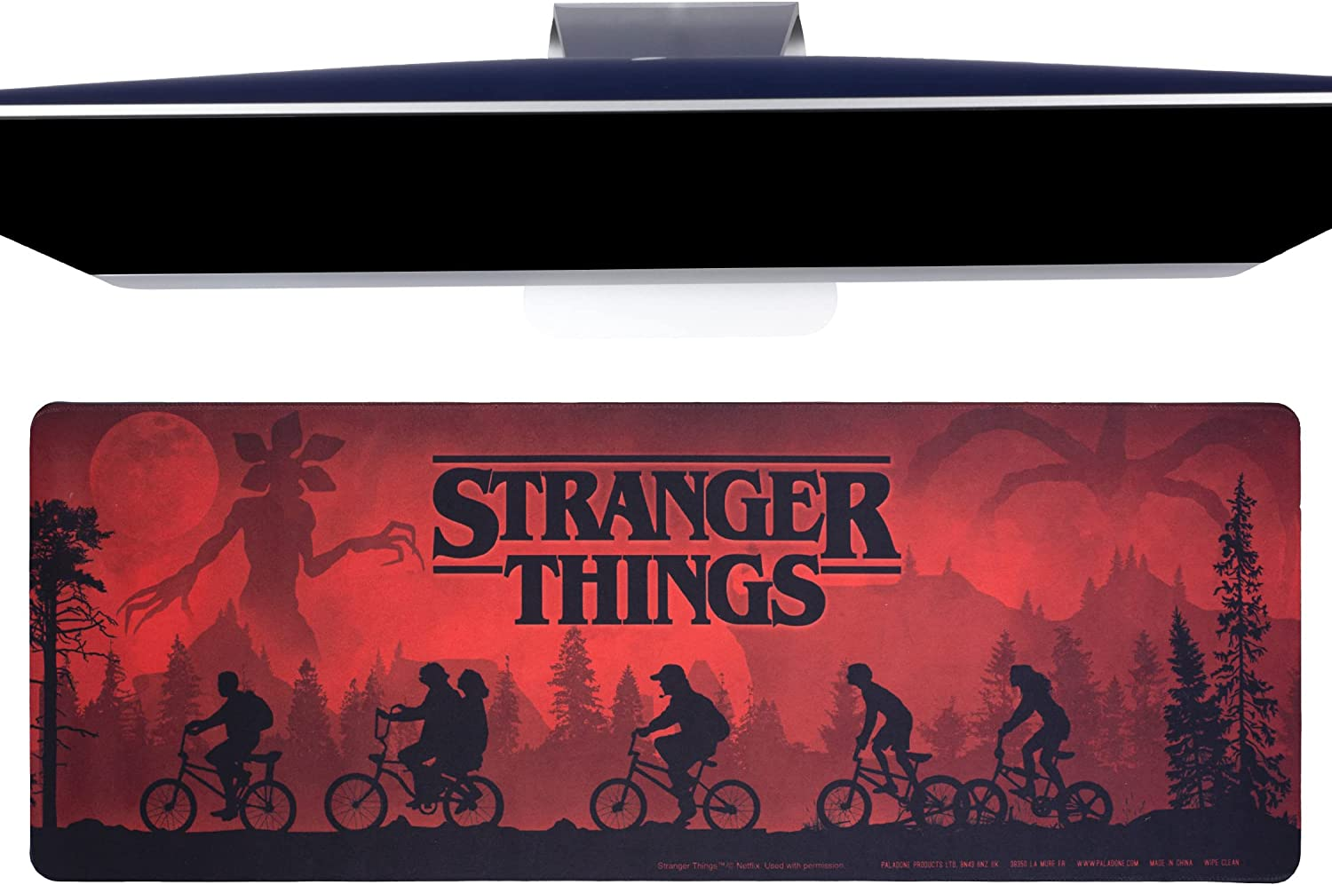 Egy számítógép monitor a Stranger Things egérpad felett helyezkedik el. A pad piros jelenetet mutat sziluett alakokkal biciklin.