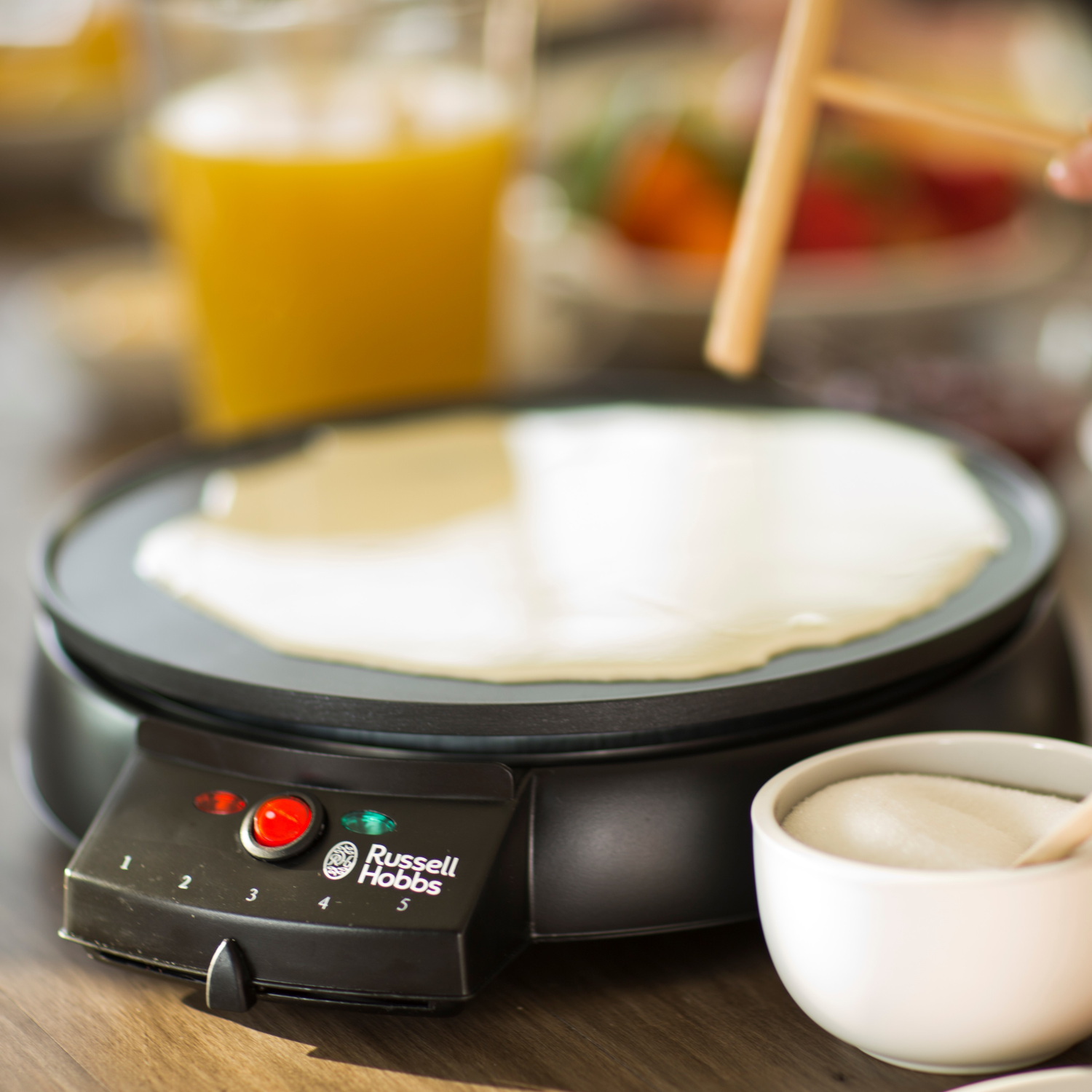 Crepe maker with crepe cooking, ingredients visible. Wooden table with orange juice and sugar.